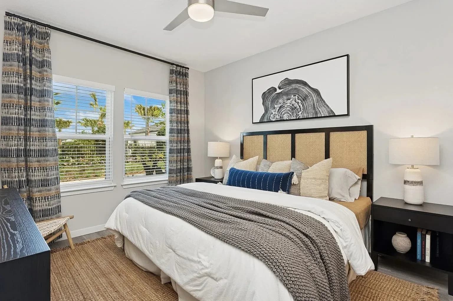 Modern bedroom with black-framed bed, woven headboard, blue and gray bedding, coastal views through windows, and patterned curtains by Jasmine Moon.