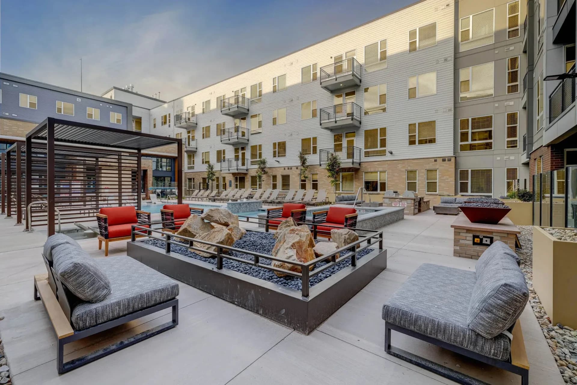 Modern courtyard with cream-colored residential building, outdoor seating with red cushions, central fire pit, and paved ground by Jasmine Moon.