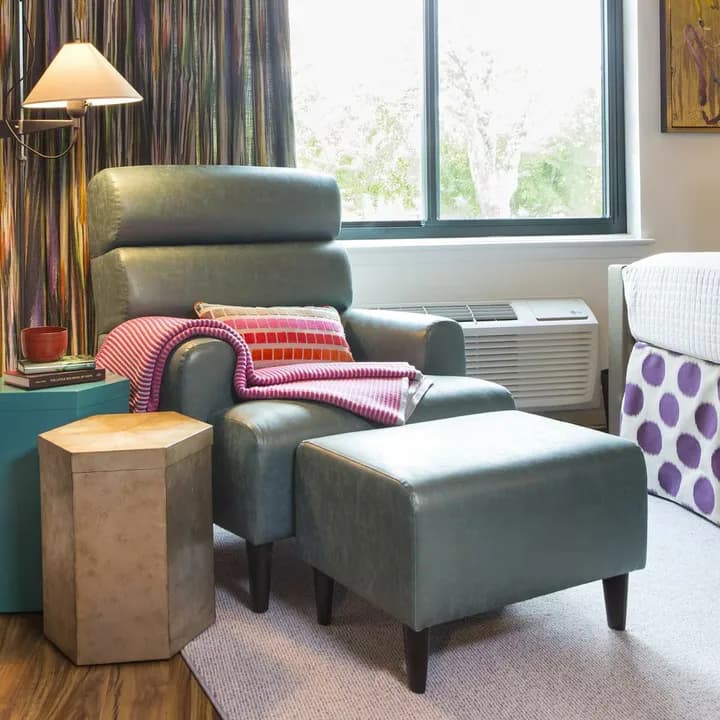 Modern living room with gray leather armchair and ottoman, wooden side table, striped curtains, and large windows overlooking greenery, designed by Jason Fox.