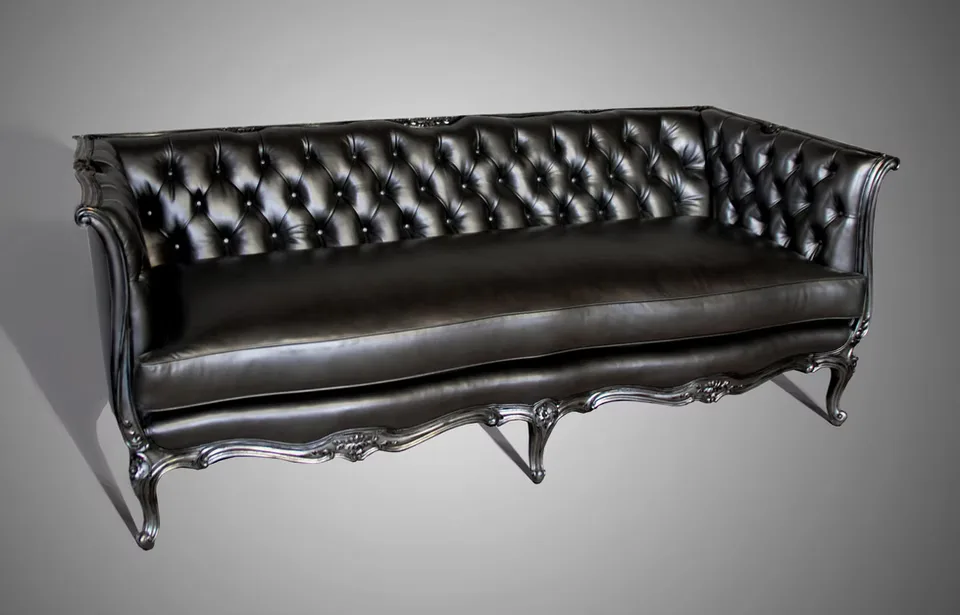 Black leather tufted sofa with ornate carved wooden legs and rolled arms against neutral background by Jason Fox.