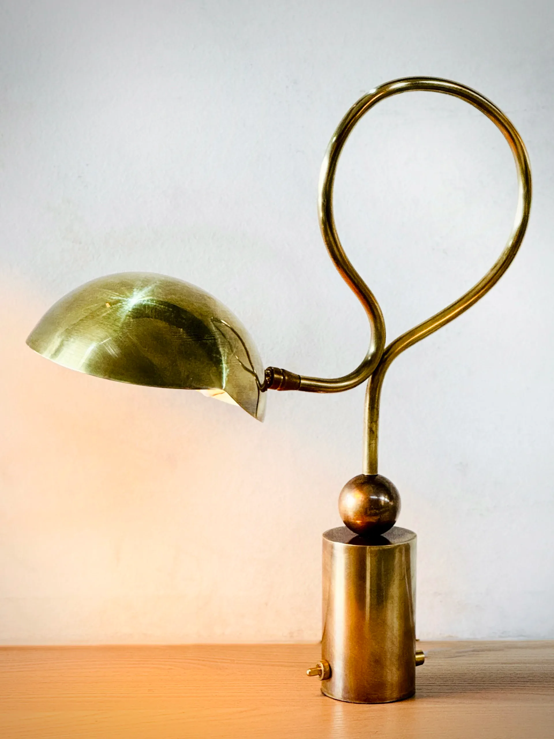 Brass sculptural table lamp with curved counterbalance arm and dome shade on wooden surface by Jason Koharik.
