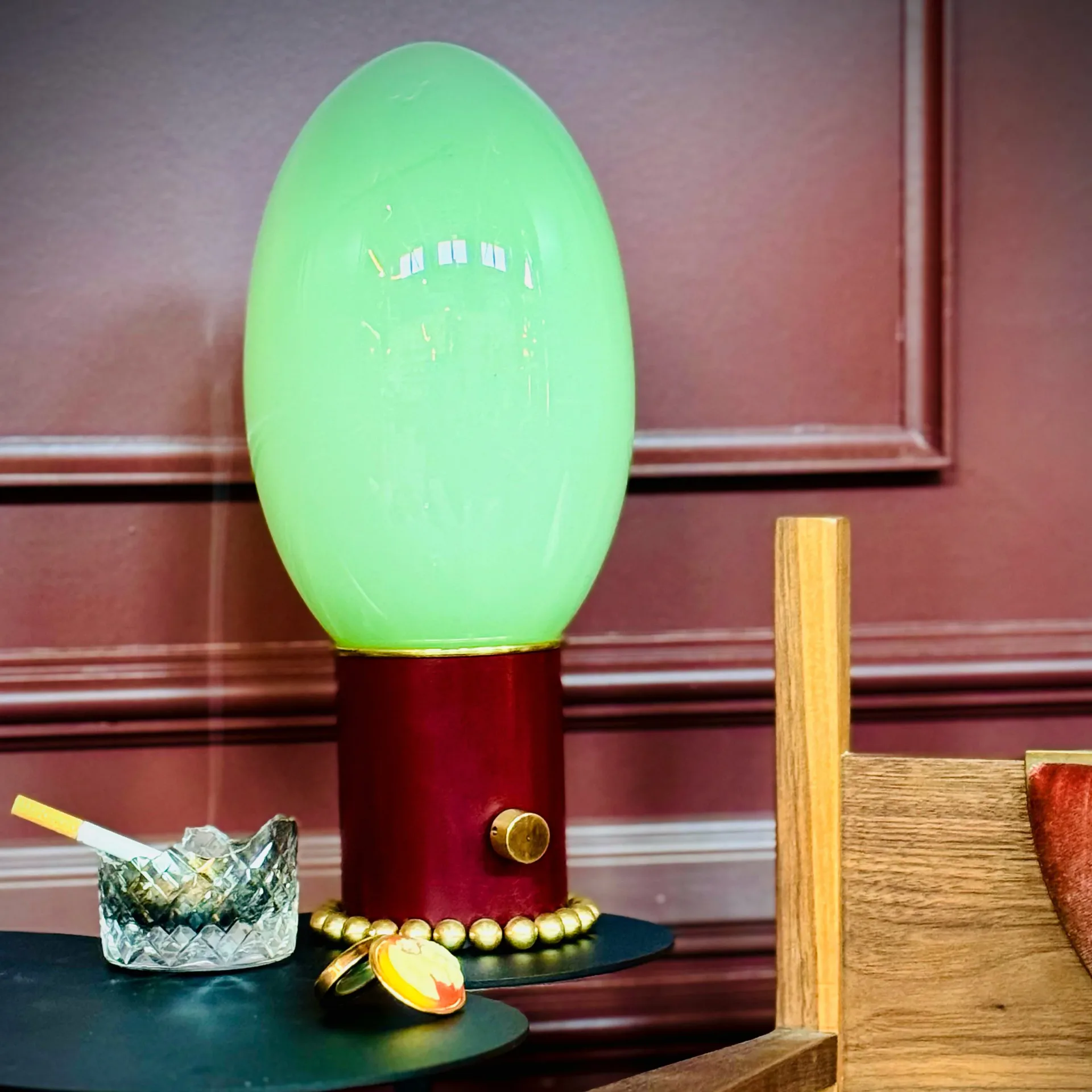 Glowing green egg-shaped lamp with burgundy base and brass beaded trim on dark teal surface, styled with crystal glass and decorative objects by Jason Koharik.