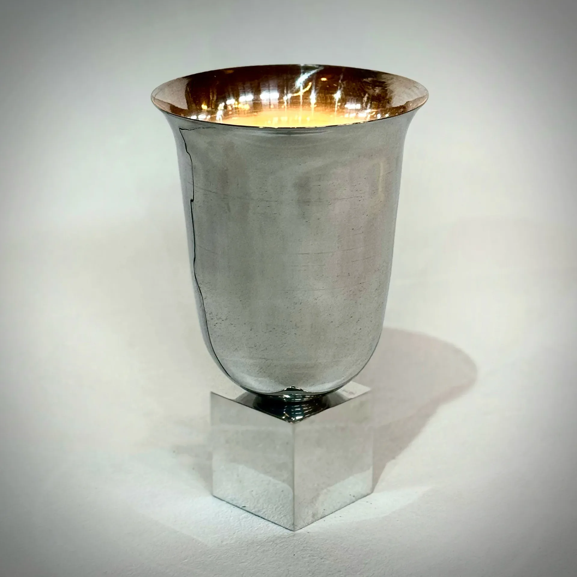 Polished metallic vessel with tapered cylindrical form, brushed silver exterior and reflective gold-toned interior, casting dramatic shadow by Jason Koharik.
