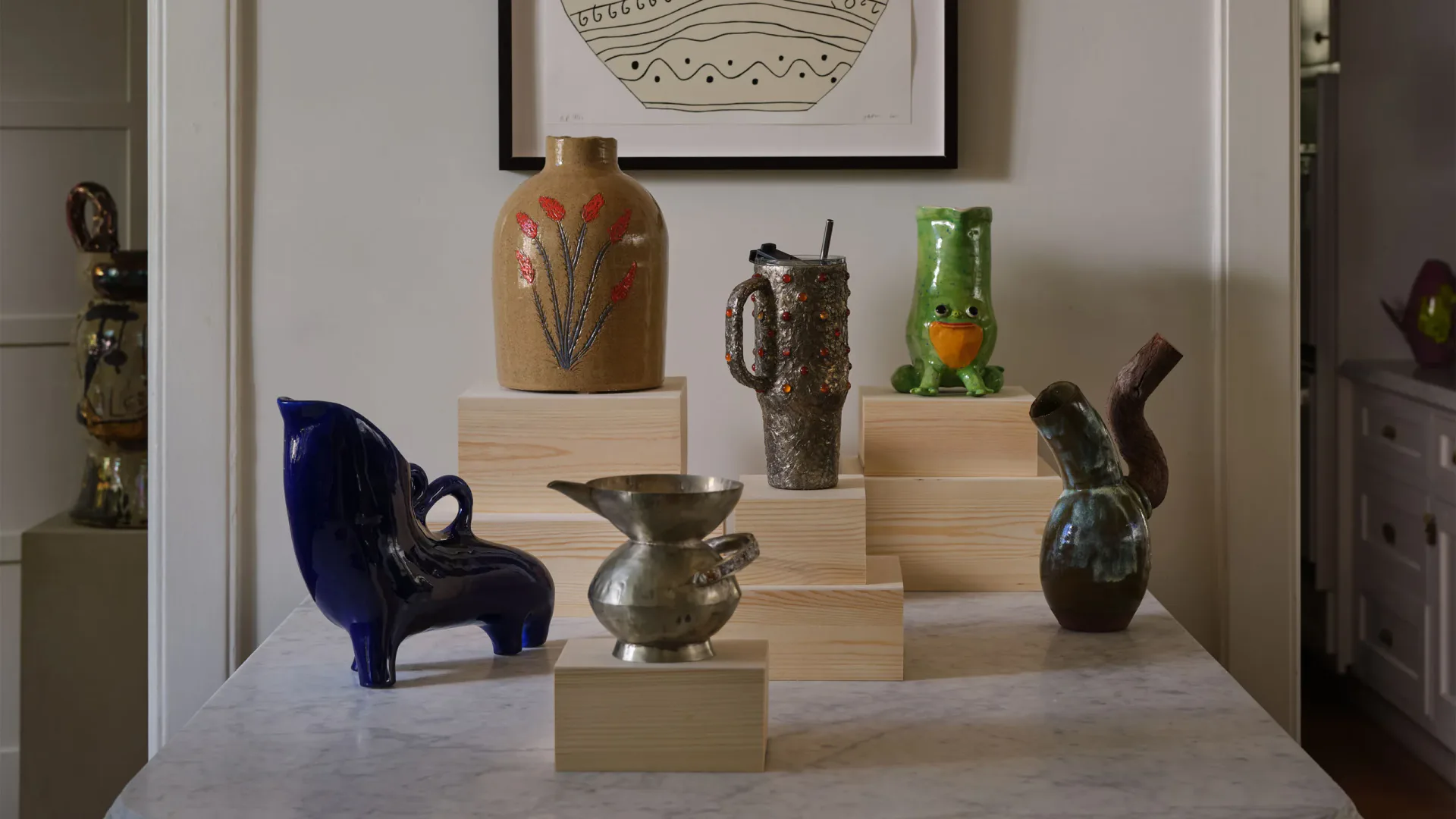 Minimalist display installation featuring wooden pedestals in varying heights with curated ceramic vessels, bronze sculptures, and decorative objects arranged against white walls by Jason Miller.