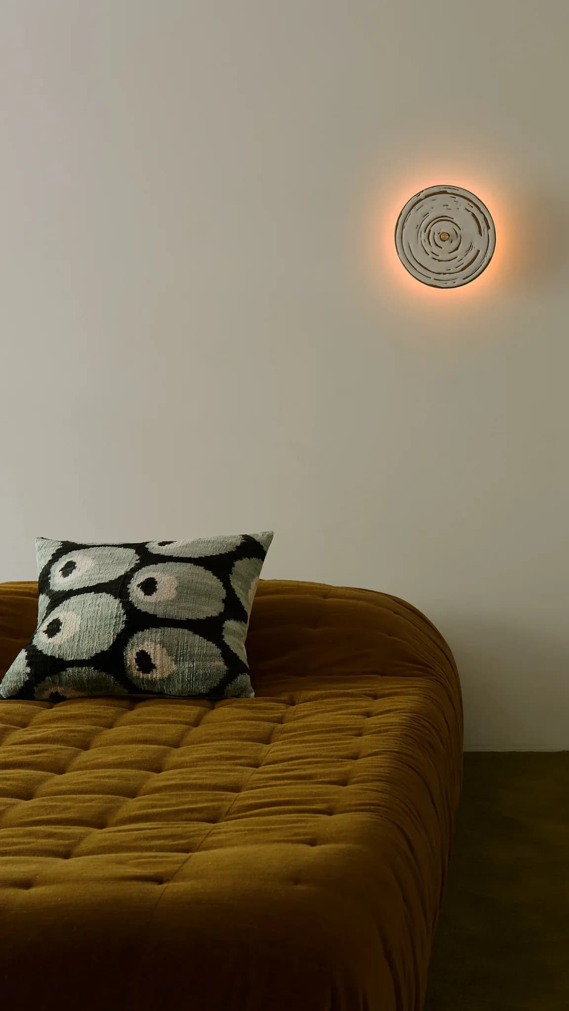 Minimalist bedroom with mustard-yellow quilted bed frame, patterned throw pillow, and circular wall-mounted lighting fixture with warm glow by Jason Miller.