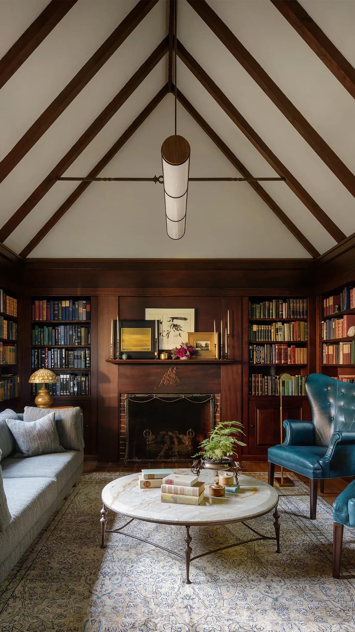 Sophisticated library lounge featuring exposed dark wood ceiling beams, built-in bookshelves, fireplace, and mid-century modern furniture including a blue leather chair and marble-top table by Jason Miller.