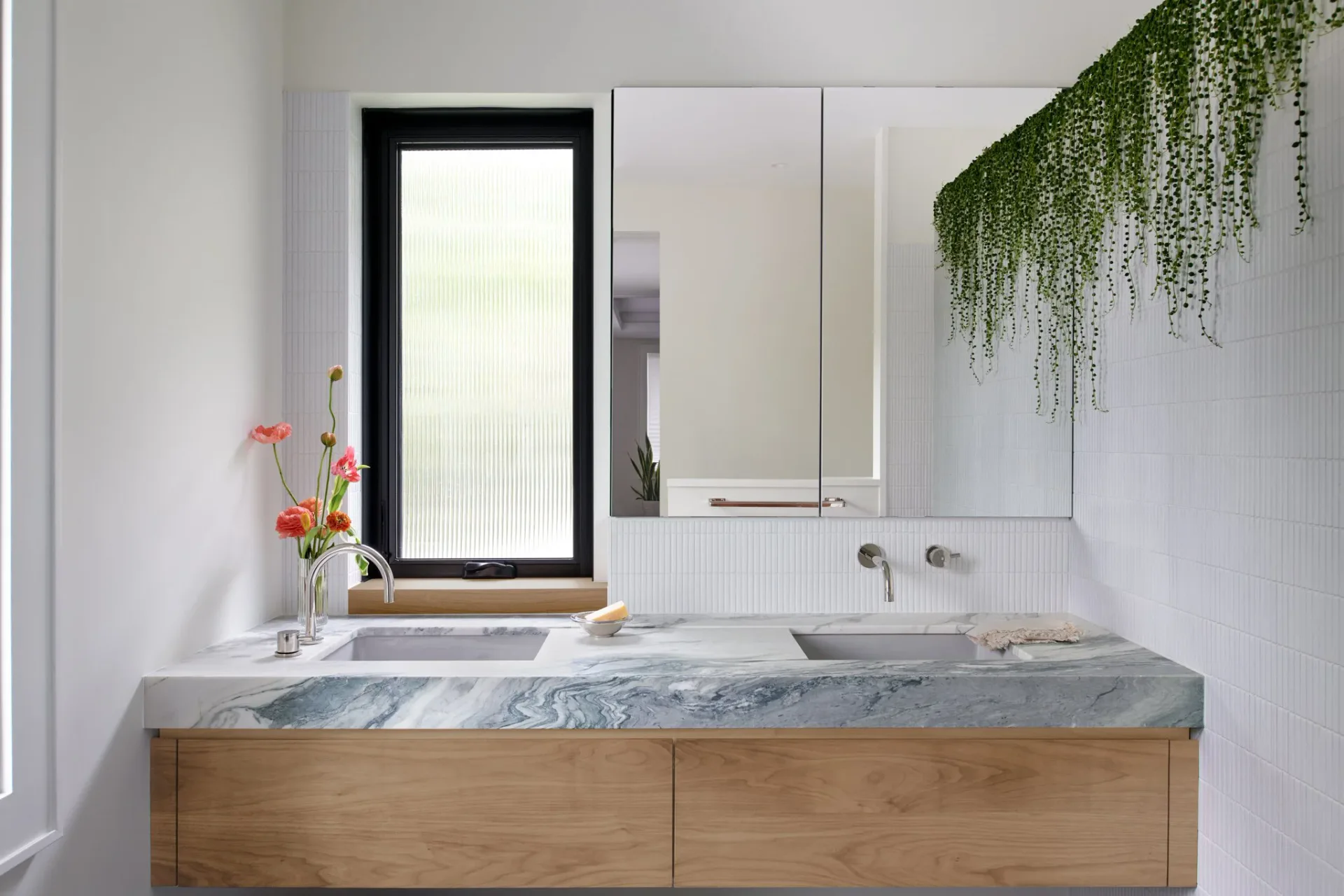 Modern bathroom vanity with marble countertop, natural wood cabinetry, and mirrored medicine cabinet, featuring a hanging plant and minimalist design by Jason Papa.