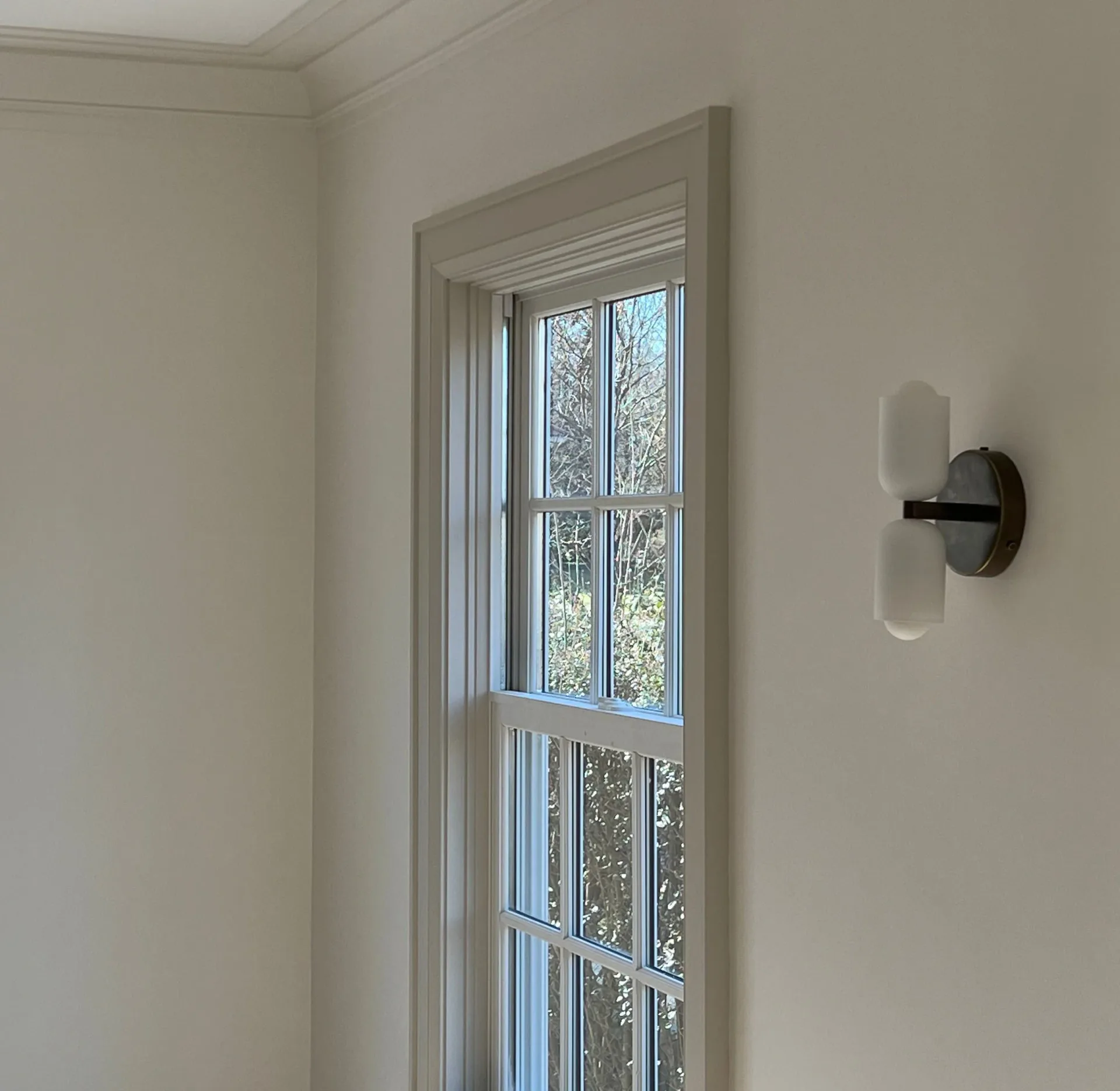 Minimalist interior with tall window framed in gray trim, white walls, and modern sconce lighting fixture by Jason Papa.