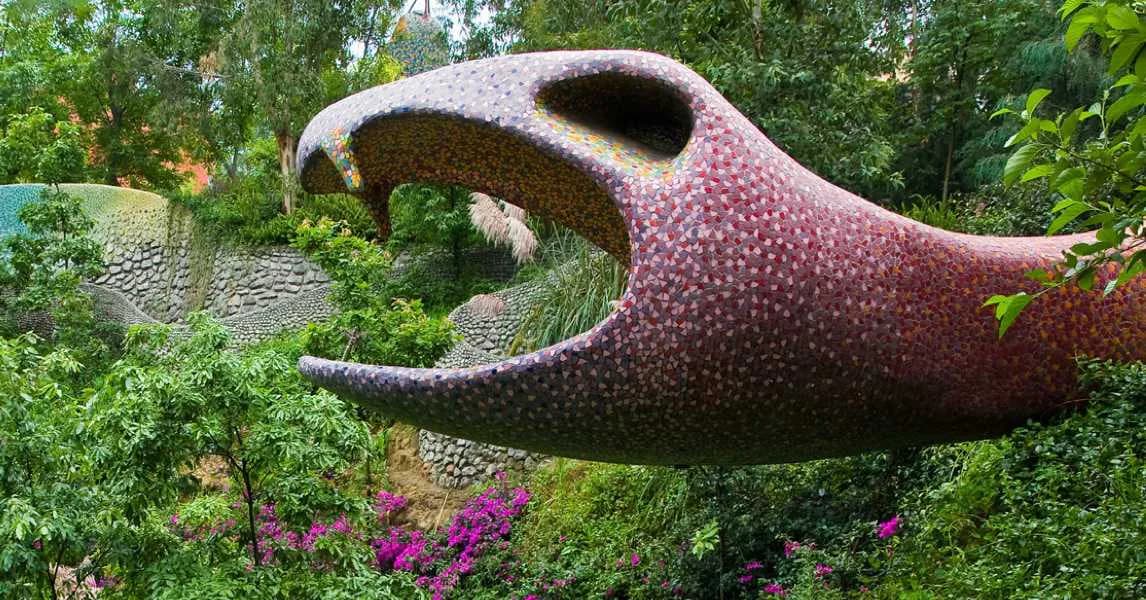 Large-scale organic sculptural installation with purple-pink textured surface and gaping mouth opening, surrounded by lush green foliage and flowering plants by Javier Senosiain.