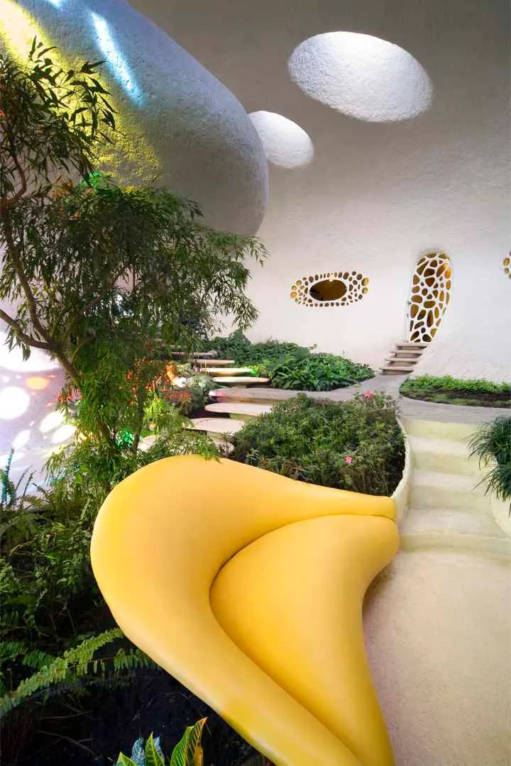 Surreal organic interior space with flowing yellow sculptural forms, layered vegetation, curved white walls with patterned openings, and floating cloud-like elements, designed by Javier Senosiain.