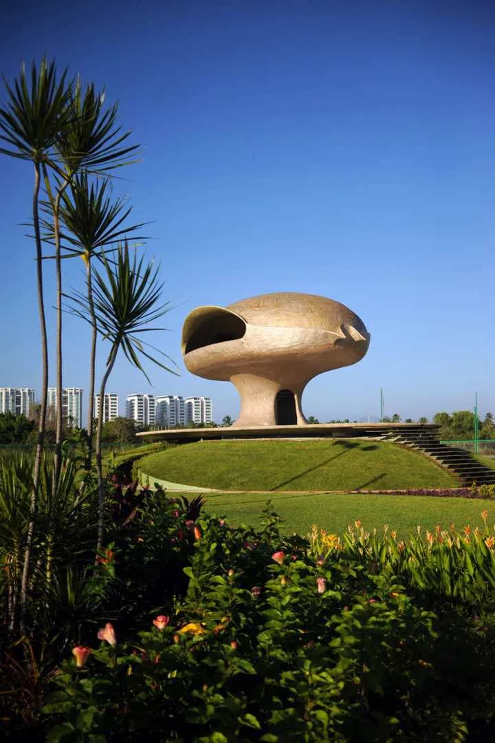 A sculptural golden architectural monument with organic, biomorphic form sits on a manicured green lawn with tropical palm trees and colorful flowering plants in foreground, by Javier Senosiain.