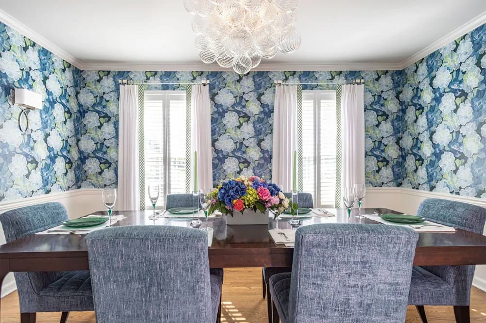 Dining room with blue floral wallpaper, white curtains, wooden table, upholstered chairs, and fresh flower centerpiece by Jean Herron.