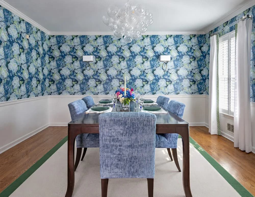 Elegant dining room with blue floral wallpaper, white wainscoting, wooden table with upholstered chairs, and glass doors by Jean Herron.