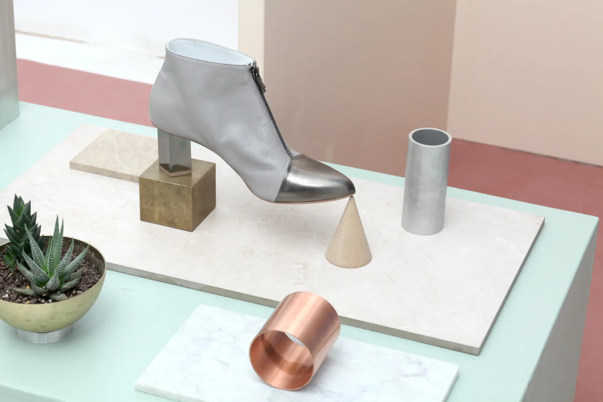 Minimalist product display featuring a gray ankle boot on geometric pedestals, metallic accessories, and potted succulent against soft pink and white backdrop by Jean Lee.