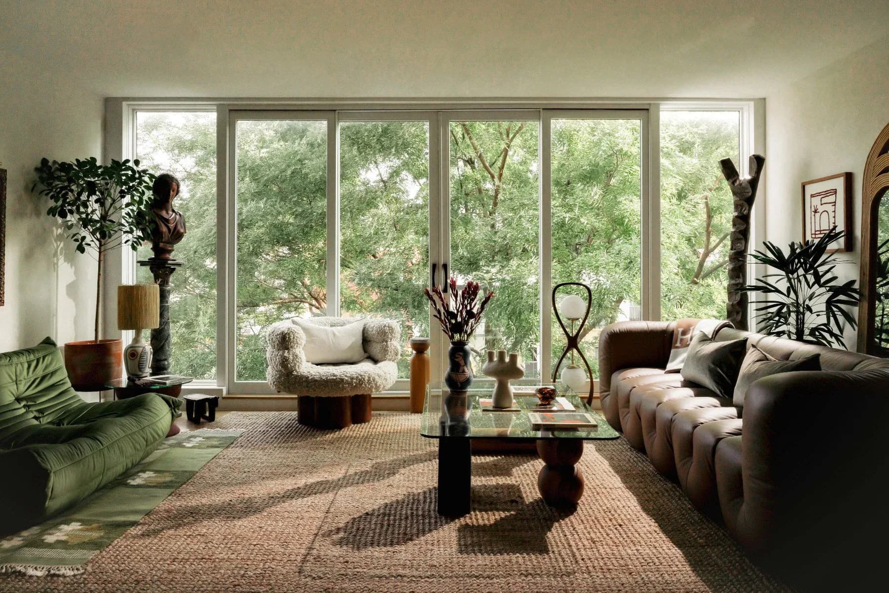 Modern living room with floor-to-ceiling windows, curved furniture, natural materials, and abundant potted plants in neutral tones designed by Jean Parker.
