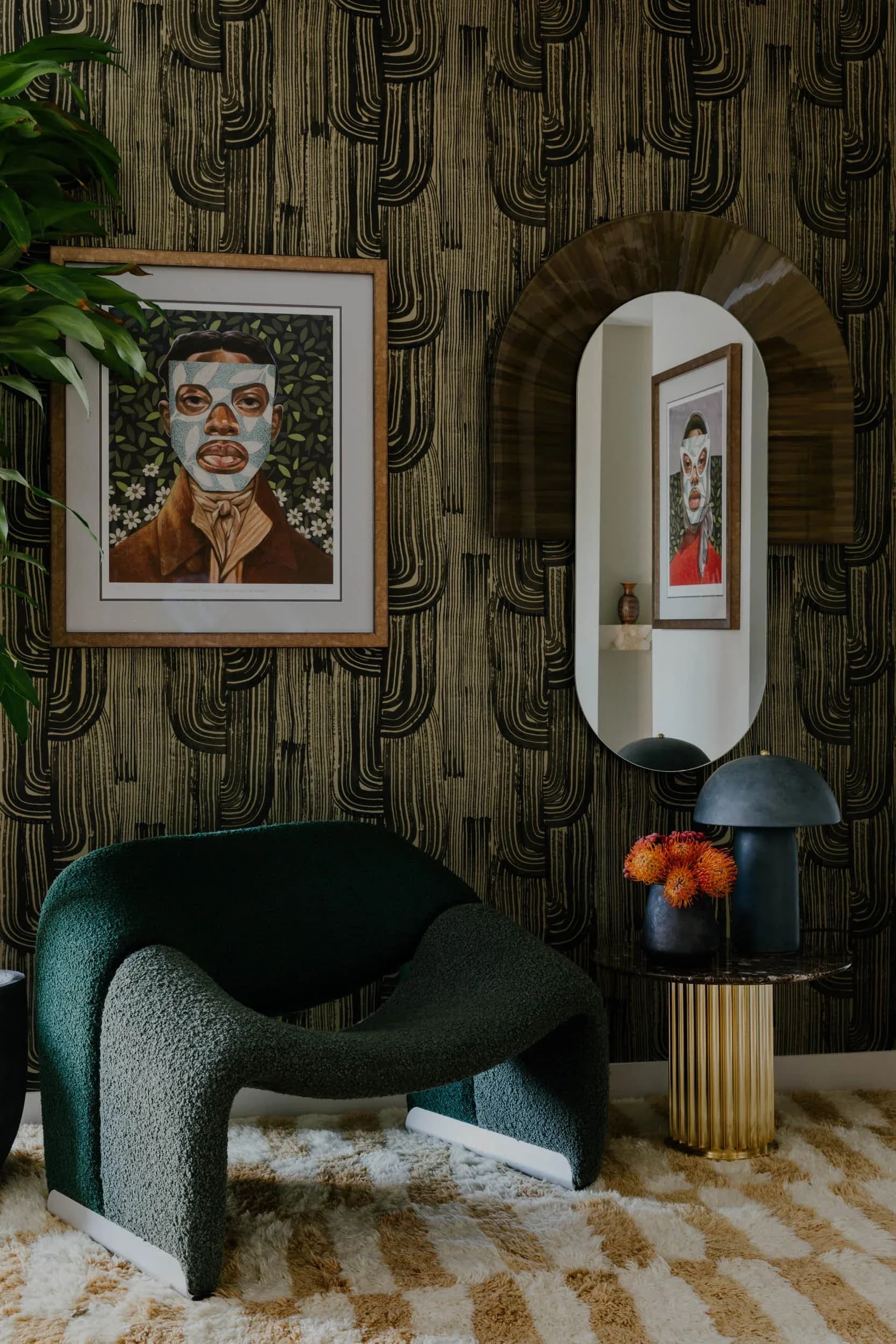 Contemporary interior featuring a curved teal armchair, patterned wallpaper, framed portrait artwork, and rounded mirror above a side table with orange flowers by Jean Parker.
