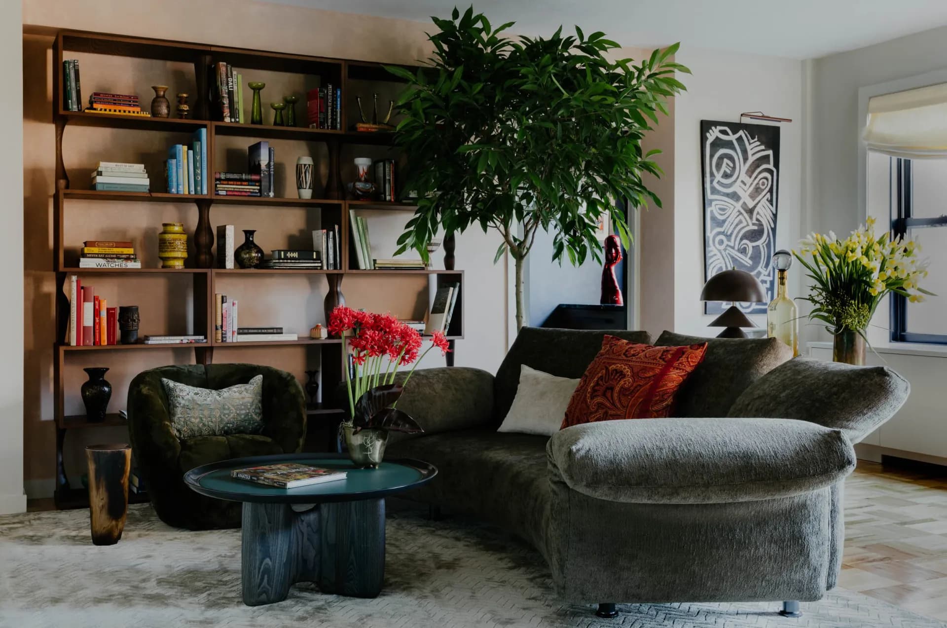 Modern living room with curved gray seating, wooden shelving unit, dark blue side table, potted greenery, and warm accent pieces in rust and coral tones by Jean Parker.