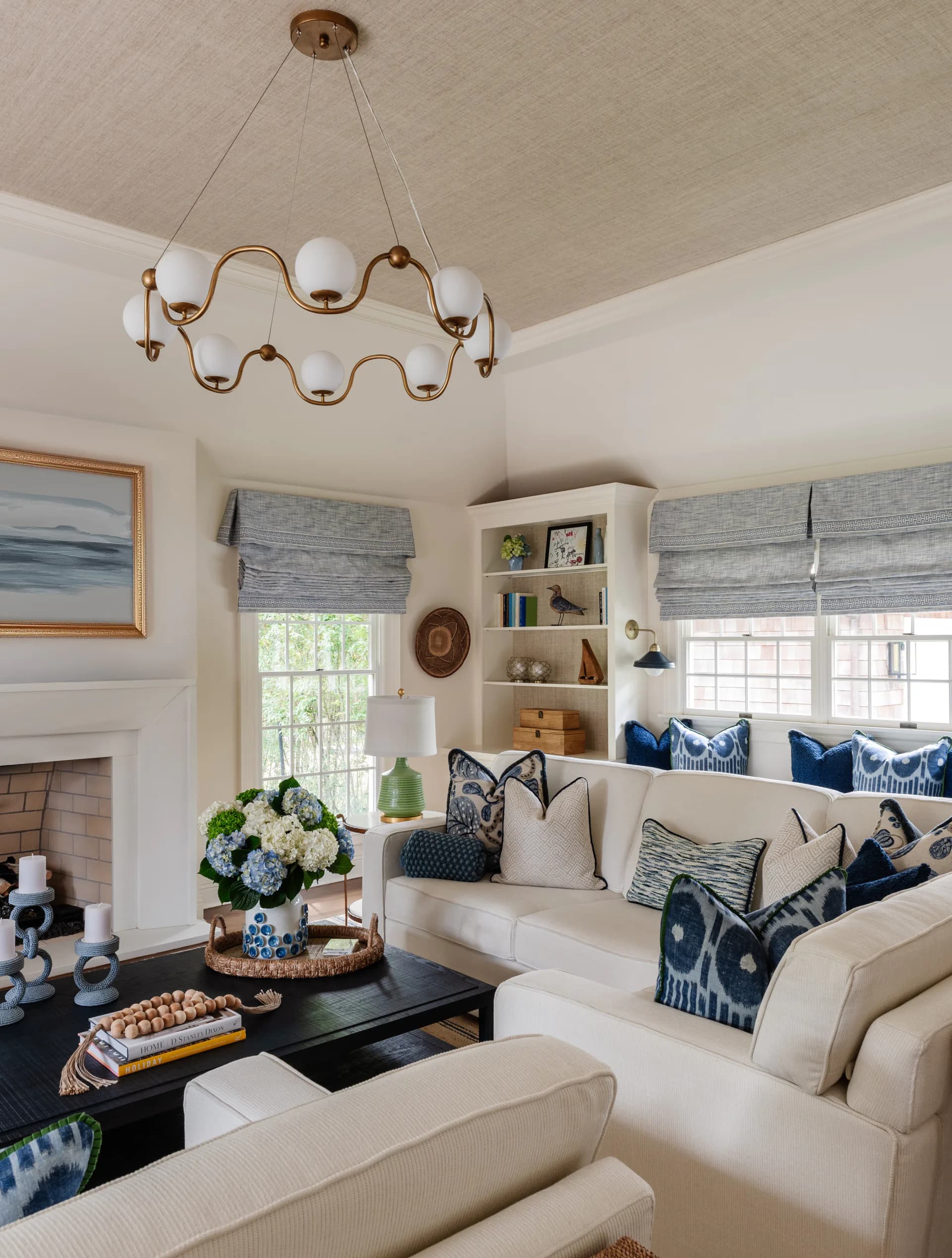 Bright coastal living room with cream upholstered seating, navy patterned pillows, brass wavy chandelier, built-in shelving, and fireplace by Jen DeLalio.