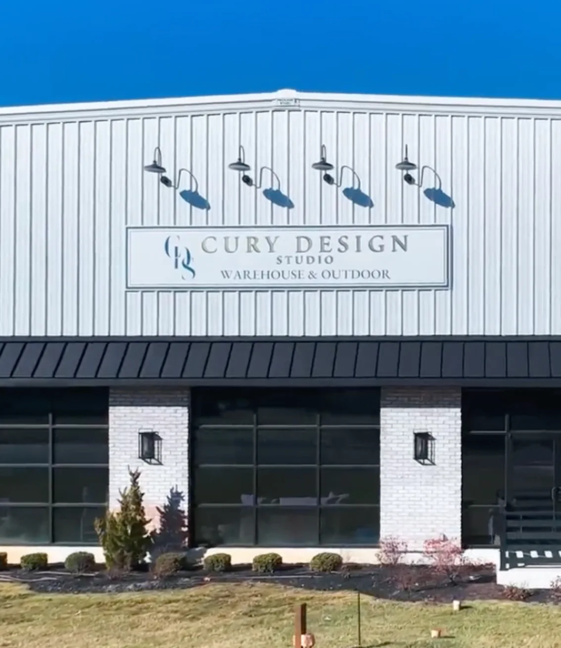 Modern warehouse storefront with white corrugated metal facade, decorative blue pendant lights, and signage for Cury Design by Jena Huynh.