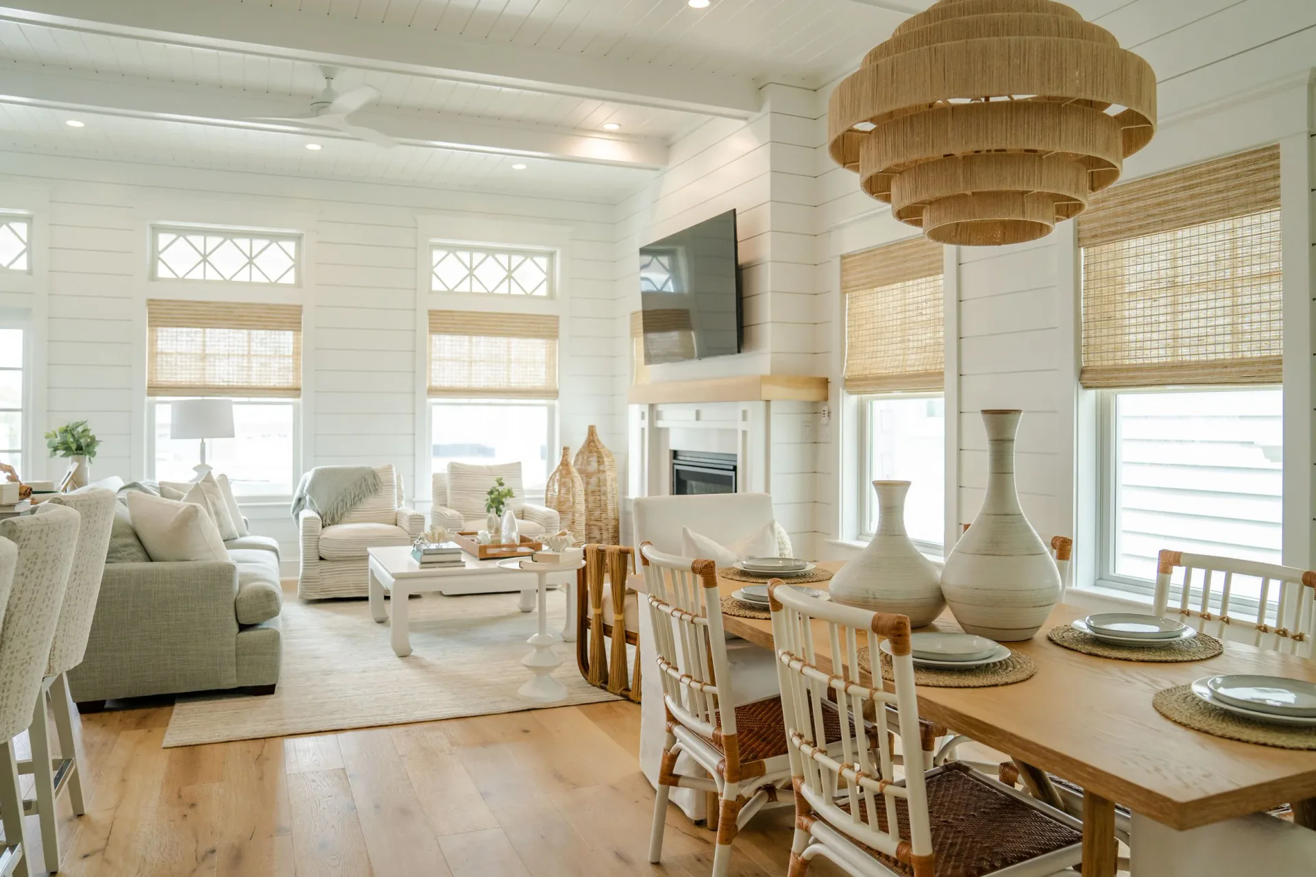 Bright open-concept living and dining space with white shiplap, natural wood furnishings, woven pendant light, and multiple windows by Jena Huynh.