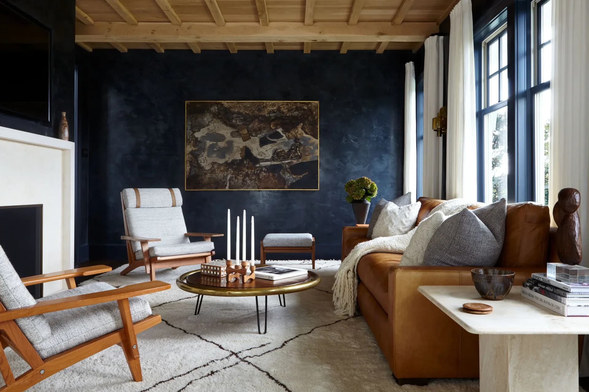 Modern living room with deep navy walls, cognac leather sofa, wooden mid-century chairs, brass coffee table, and gold-framed artwork by Jena White.