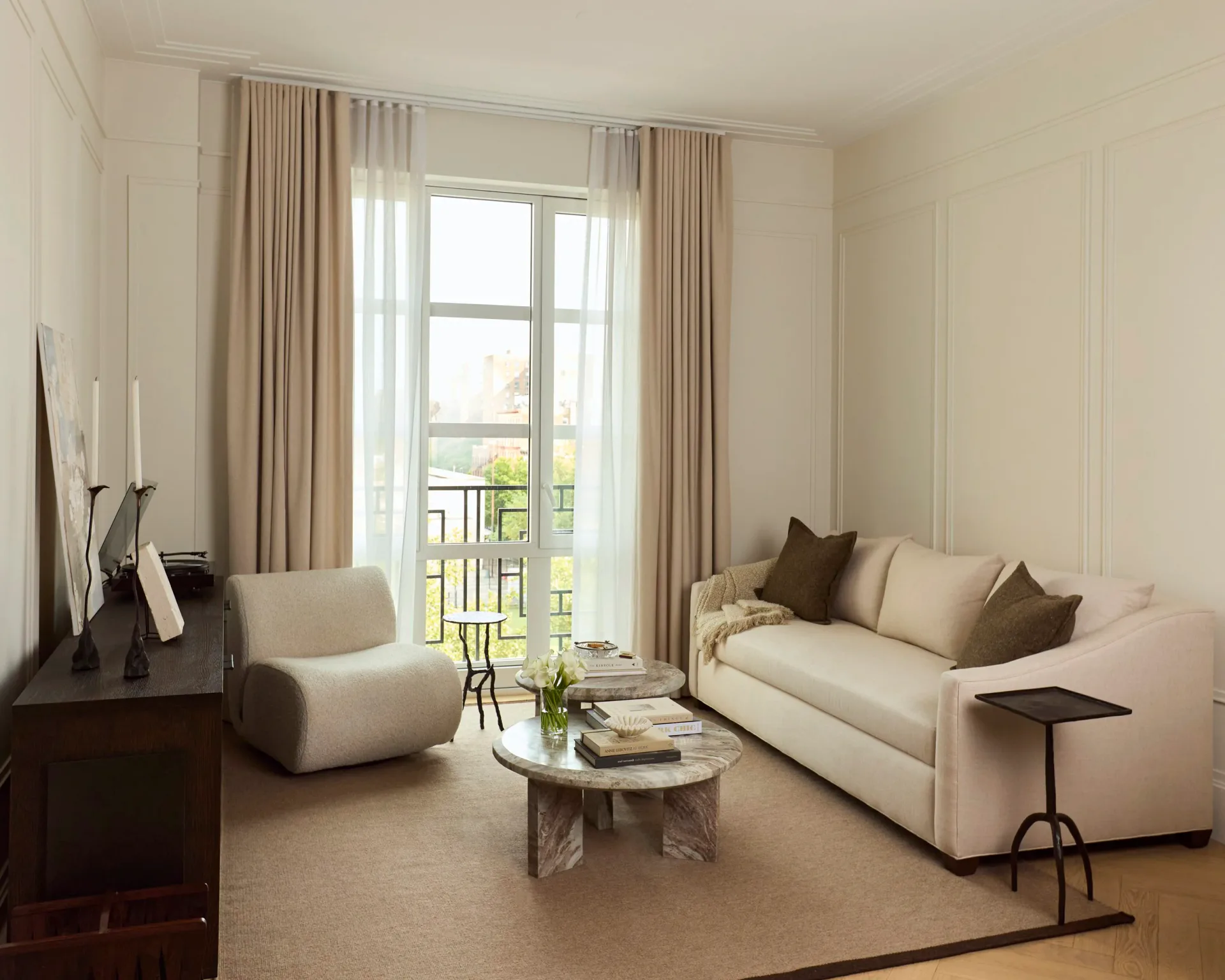 Bright contemporary living room with cream sofa, neutral armchair, glass coffee table, and floor-to-ceiling windows with elegant drapery, designed by Jenna Reinero.