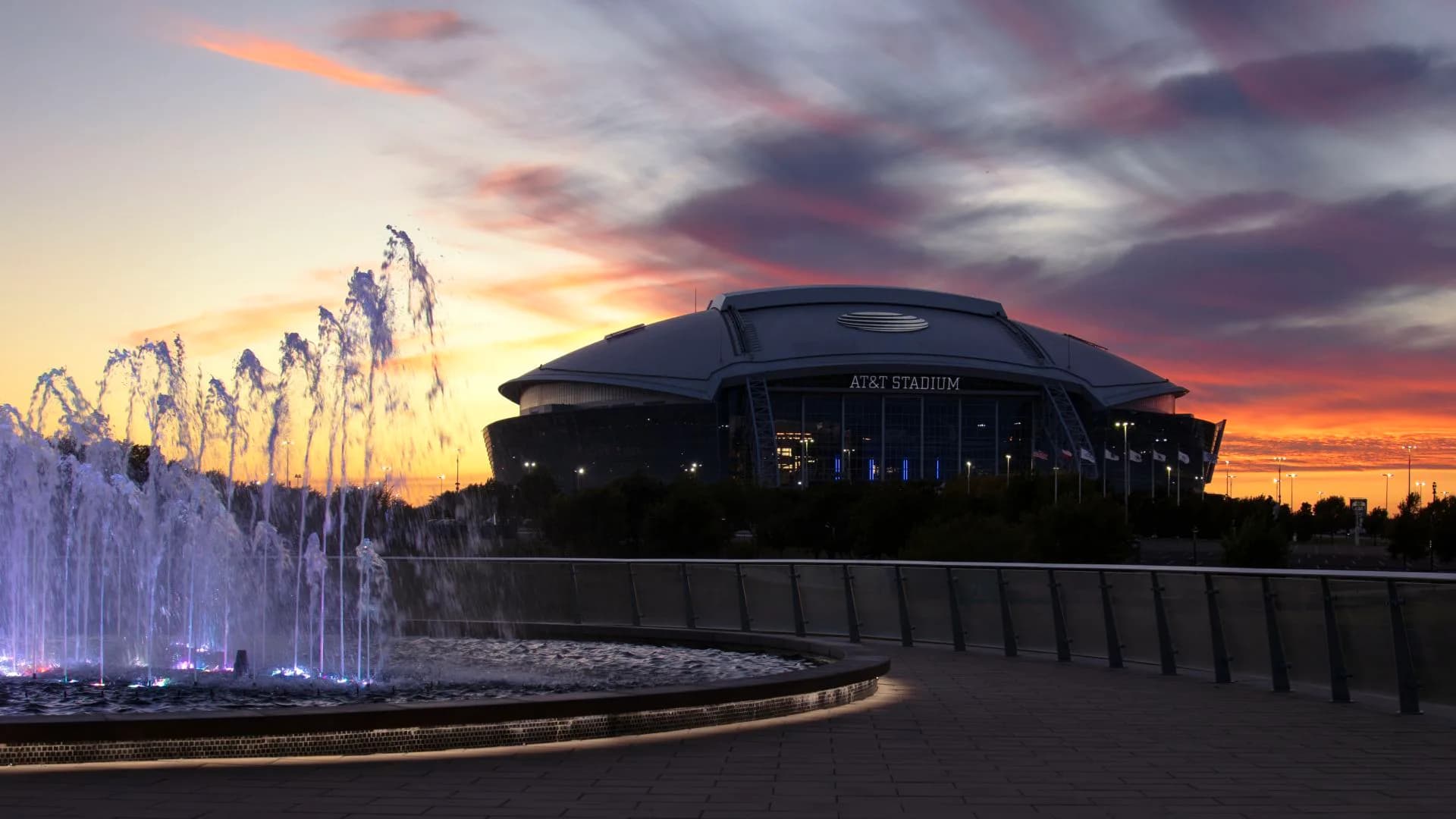 Modern arena building with dark curved architecture at sunset, illuminated fountain in foreground, dramatic sky, by Jennifer Mehra.