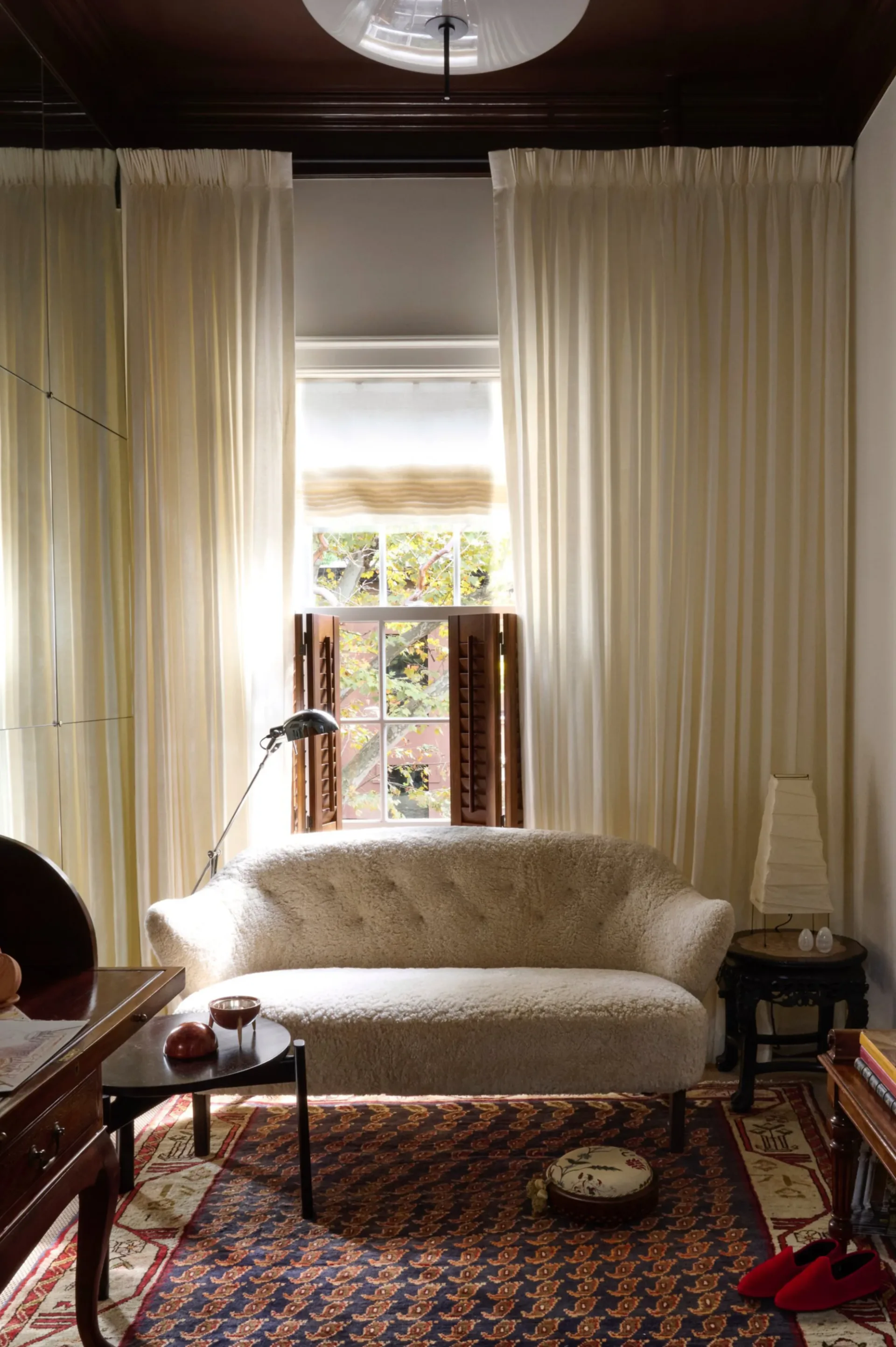 Cream curved sofa positioned beneath tall windows framed by flowing cream curtains, with patterned rug and dark wood furniture in a light-filled room by Jennifer Paccione Angulo.