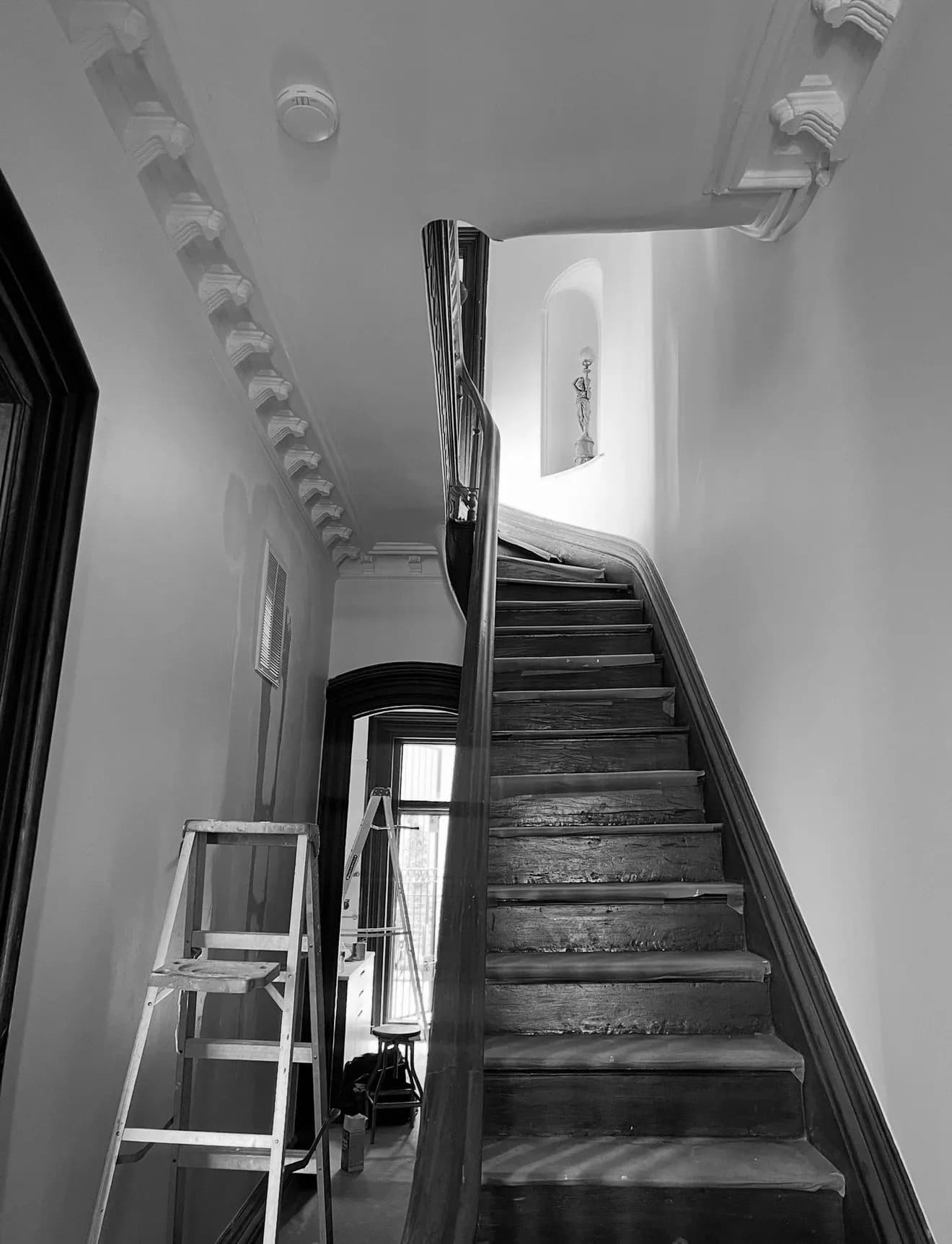 Black and white stairwell with dark wooden stairs, metal railings, and white walls lit by natural light from above, by Jennifer Paccione Angulo.