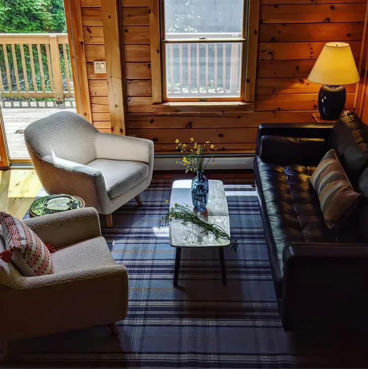 Modern cabin living room with wood walls, plaid area rug, gray upholstered chairs, black sofa, and glass coffee table overlooking a deck by Jennifer Salvemini.