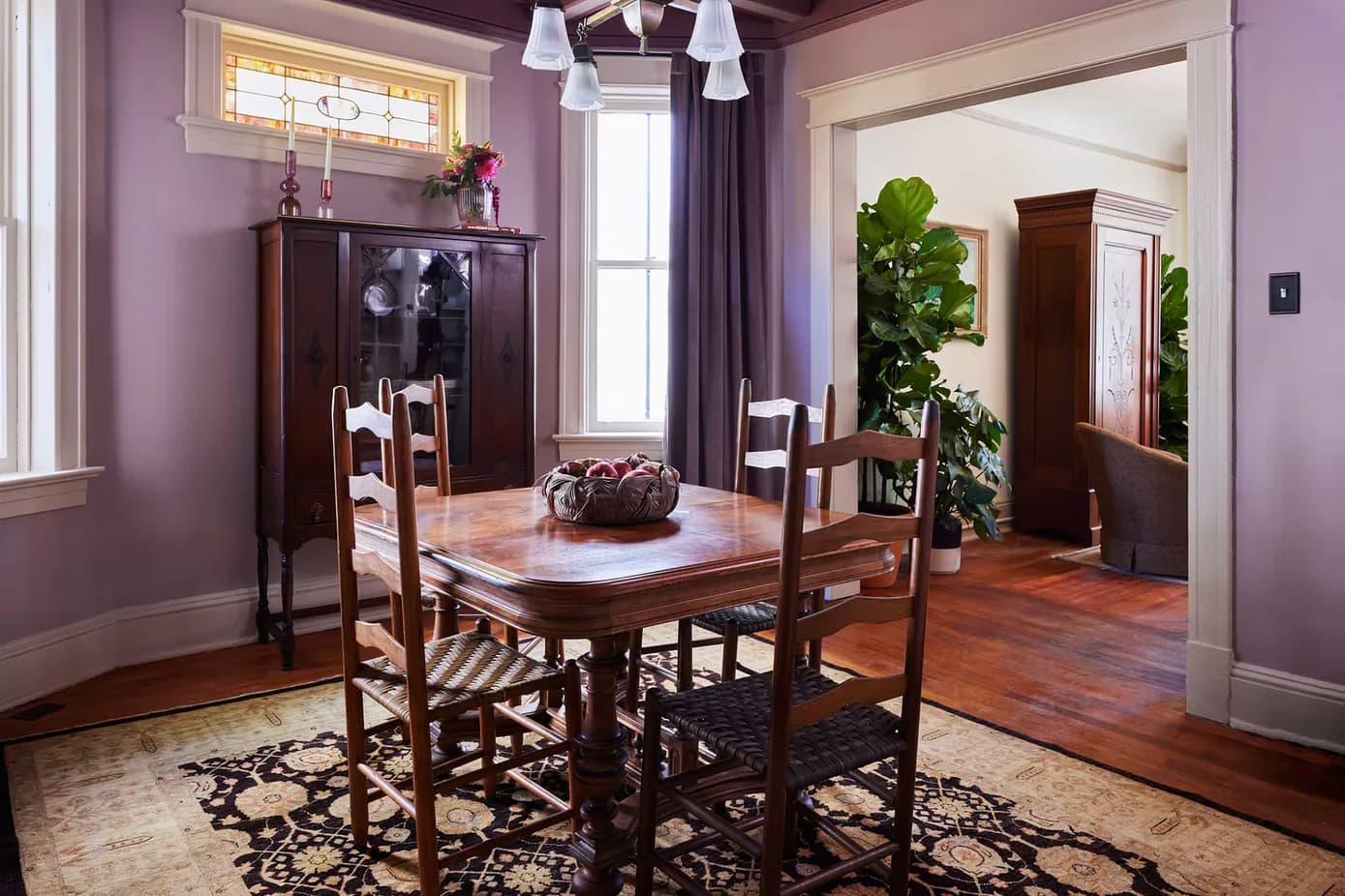 Purple dining room with round wood table, rush-seat chairs, dark china cabinet, patterned area rug, and potted plants by Jennifer Salvemini.