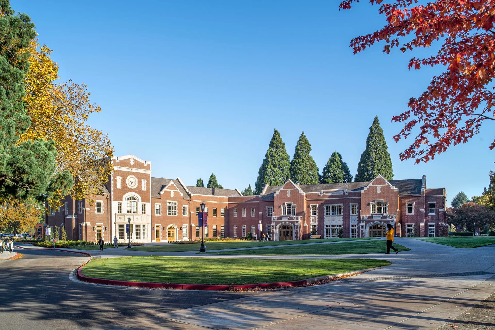 Brick institutional buildings with classical architecture arranged around a manicured lawn, featuring warm autumn foliage and clear blue sky, by Jennifer Stone.