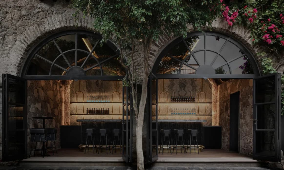 Symmetrical wine bar interior with arched brick alcoves, warm wood cabinetry, black metal doors, and climbing ivy above by Jennifer Tsai.