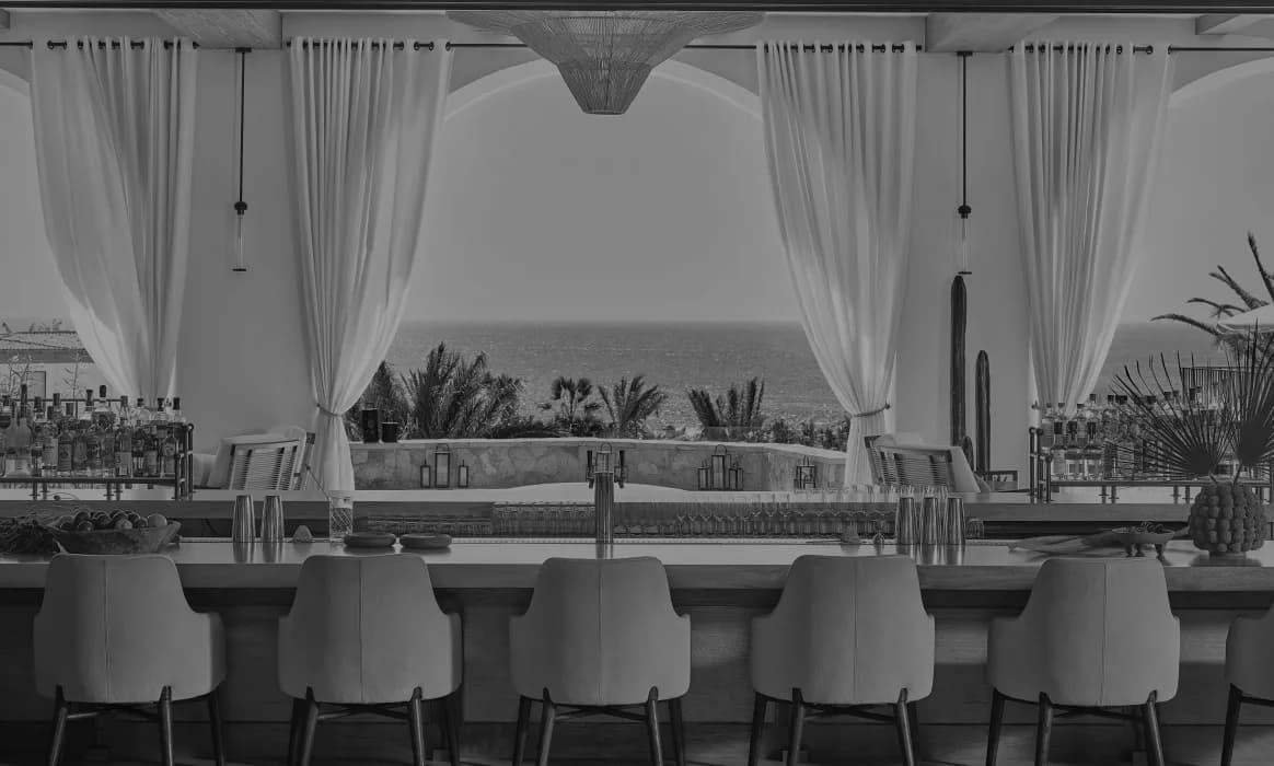 Modern coastal bar interior with white barrel seating, sheer curtains, and ocean views, designed by Jennifer Tsai.
