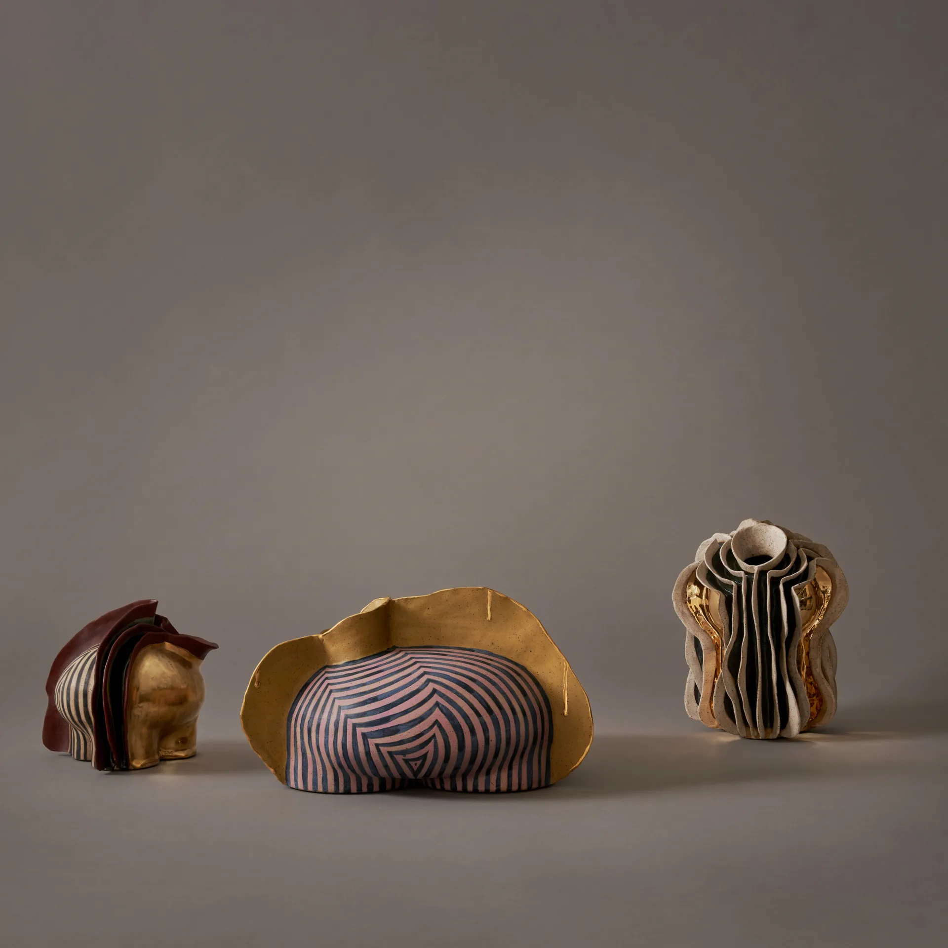 Three sculptural objects with gold and striped ceramic elements arranged on a neutral surface, showcasing organic forms and mixed materials by Jeremy Anderson.