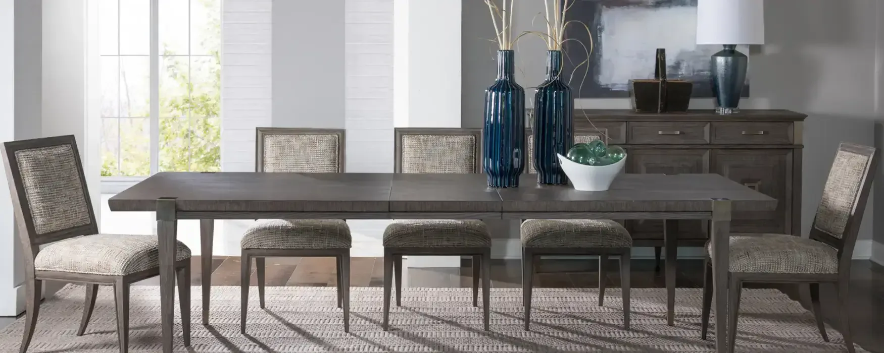 Modern dining room with gray wood table, upholstered chairs, and neutral palette featuring decorative blue vessels and sideboard by Jerry Rock.