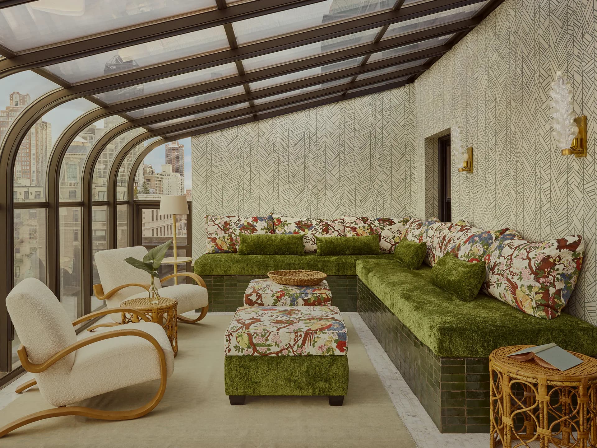 A sunroom with curved glass ceiling features built-in green velvet seating, floral patterned cushions, and mid-century modern accents by Jess Nahon.