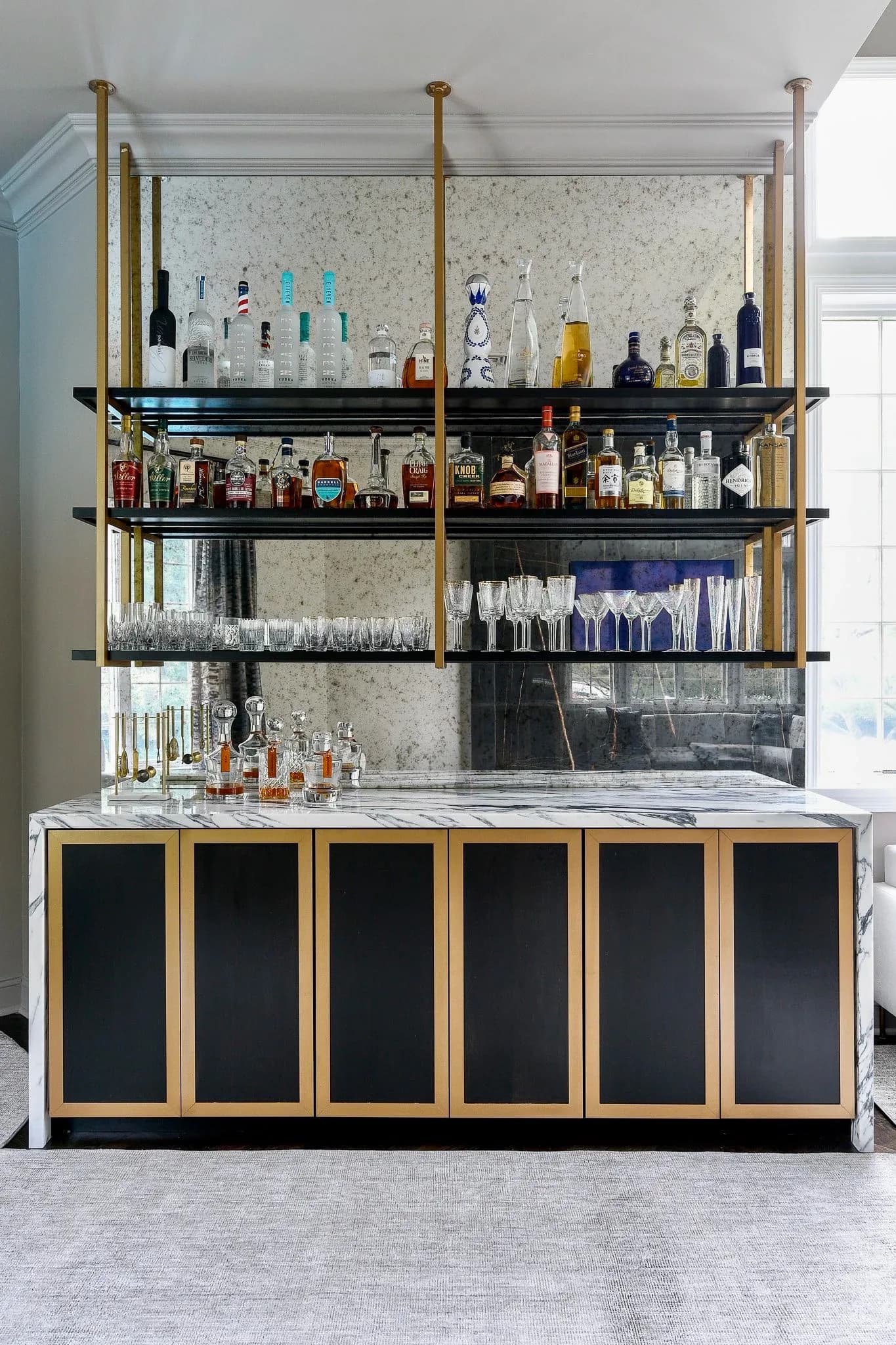 A modern bar interior with brass-framed shelving displaying spirits and glassware above a marble-topped counter with black cabinetry by Jessica Cohen.