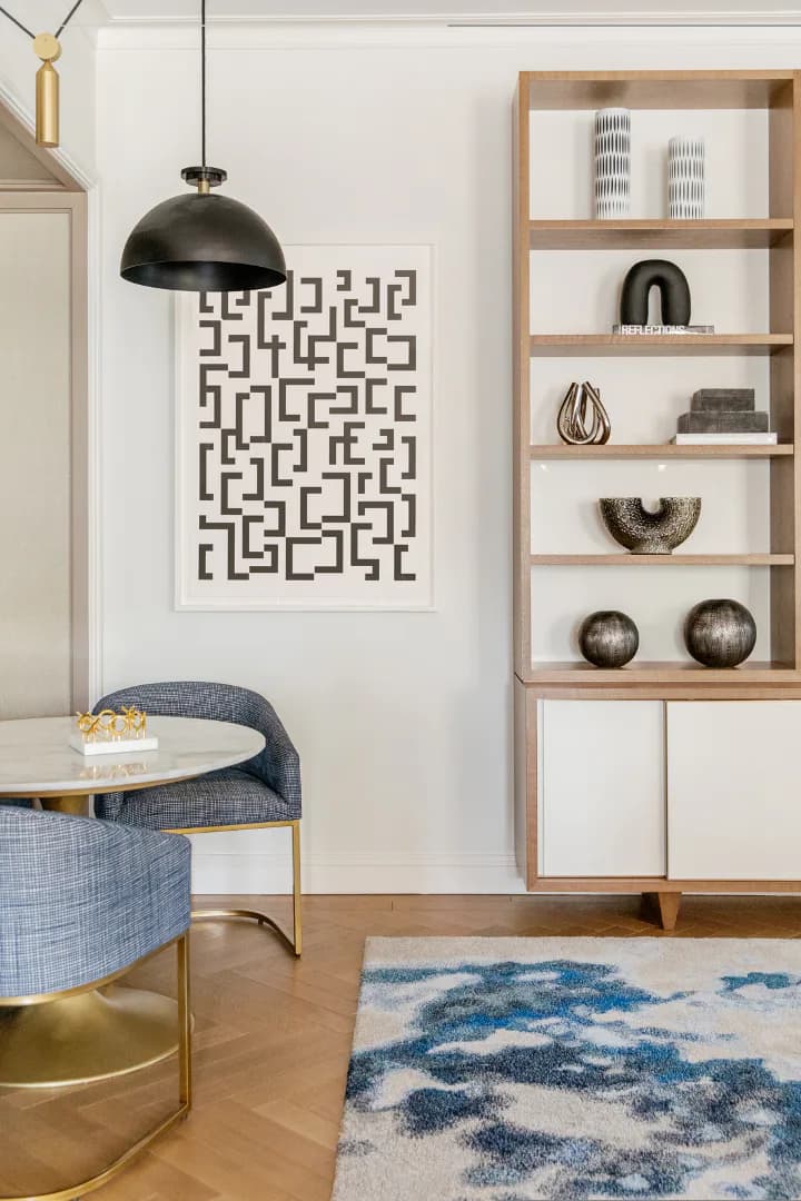 Modern living room with blue upholstered chair, natural wood shelving unit, black pendant light, geometric wall art, and patterned blue area rug by Jessica Cohen.