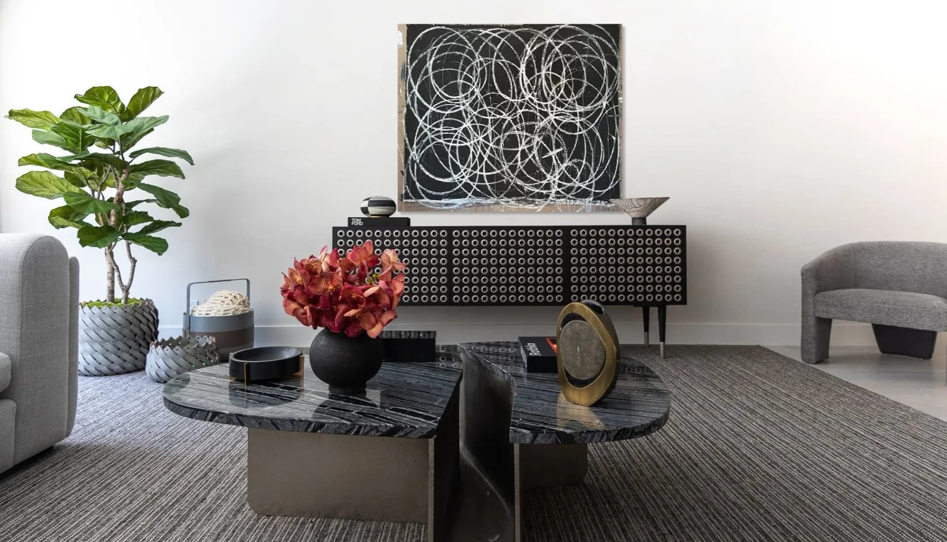 Modern living room with black and grey palette featuring curved furniture, sculptural black tables, artwork, plants, and geometric wall installation by Jessica Cohen.