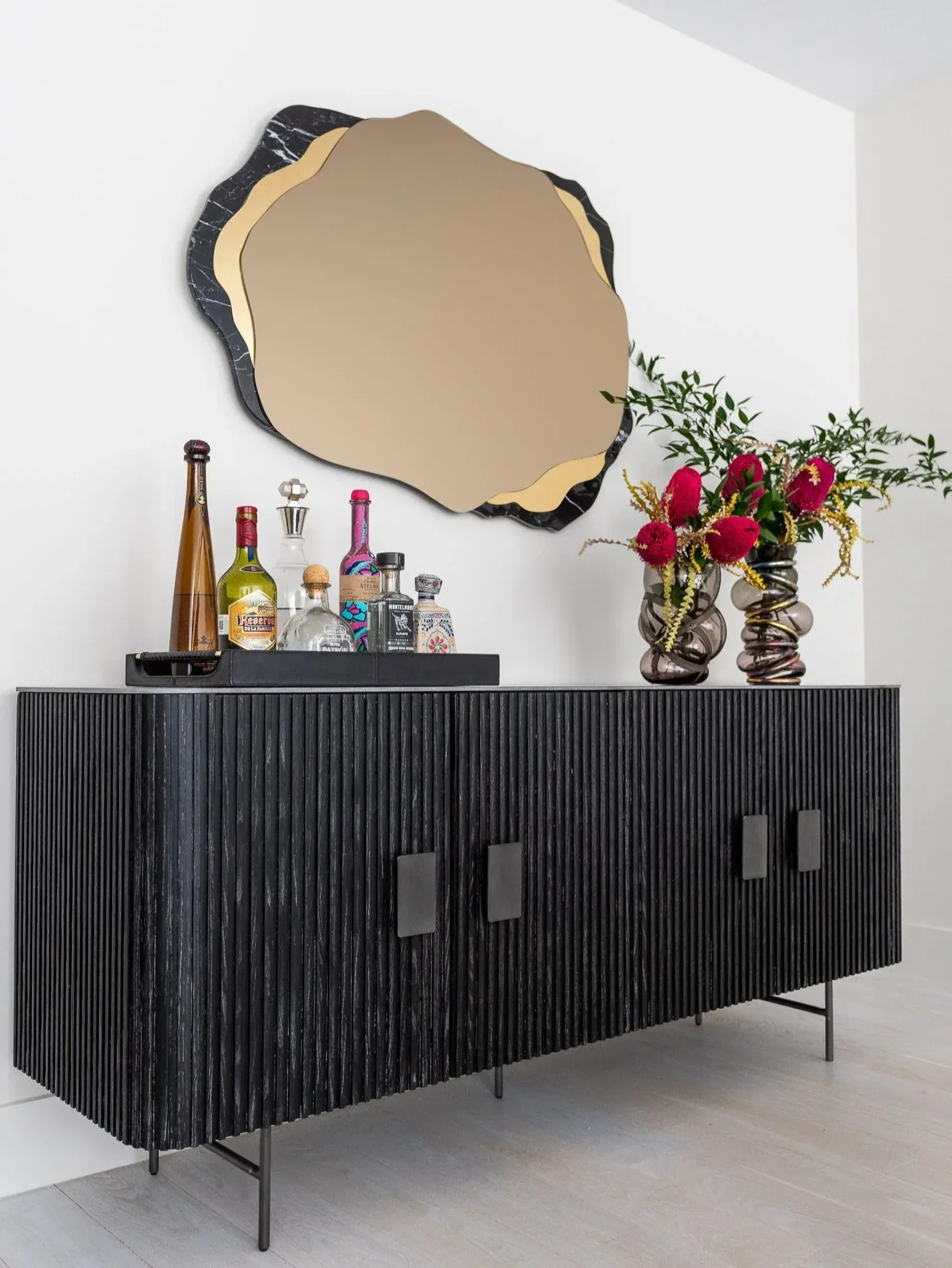 Modern black ribbed credenza with metal legs styled with champagne bottles, perfumes, and a vibrant red floral arrangement beneath an organic-shaped mirror by Jessica Cohen.