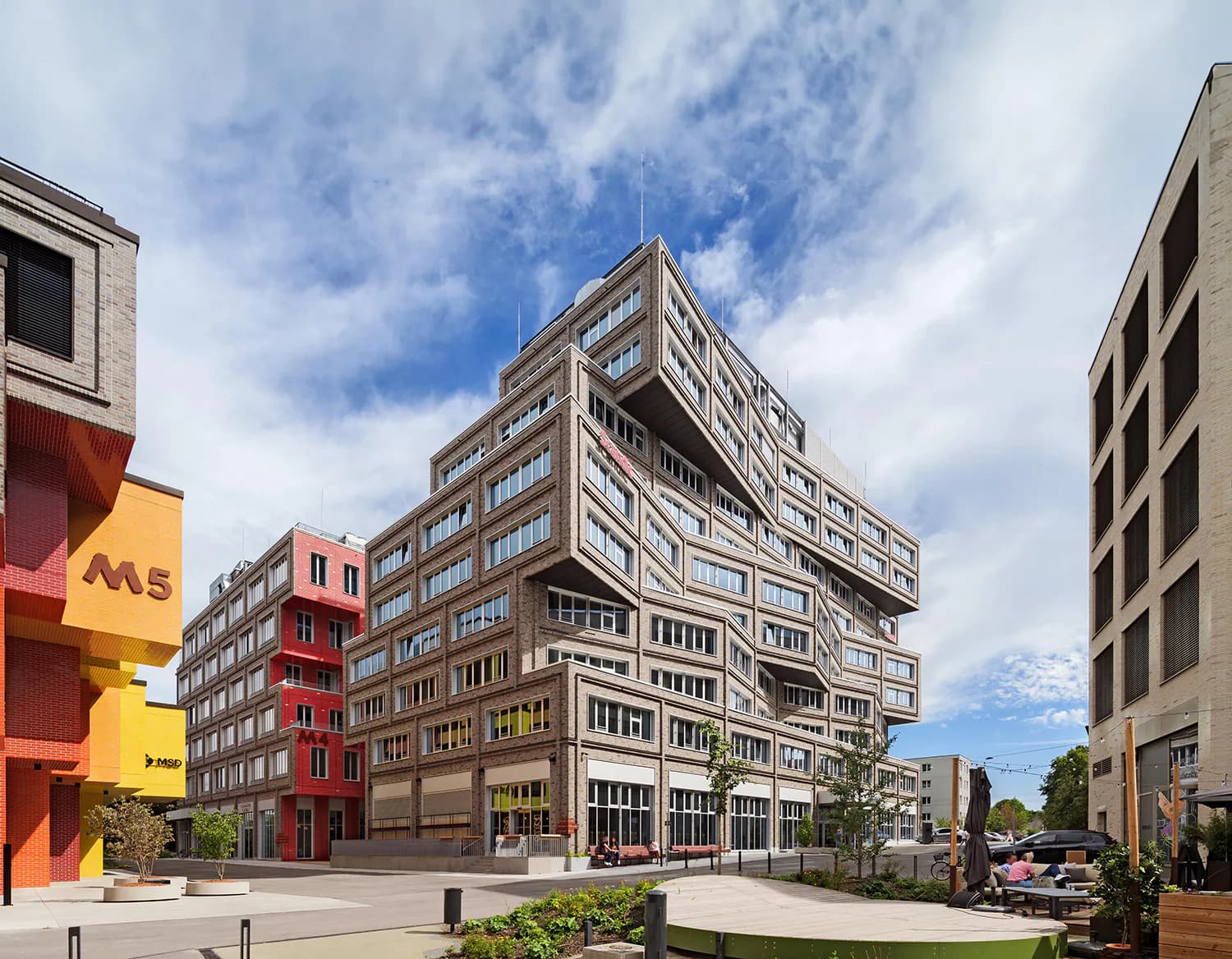 Contemporary mixed-use building with stacked residential volumes and ground-floor retail, featuring warm-toned brick and modern window treatments in an urban plaza setting by Jessica Knobloch-Arscott.
