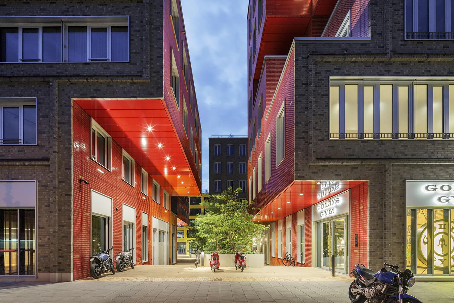 Evening courtyard with red-orange modern canopy structures, brick buildings, illuminated storefronts, and pedestrians in a contemporary urban plaza by Jessica Knobloch-Arscott.