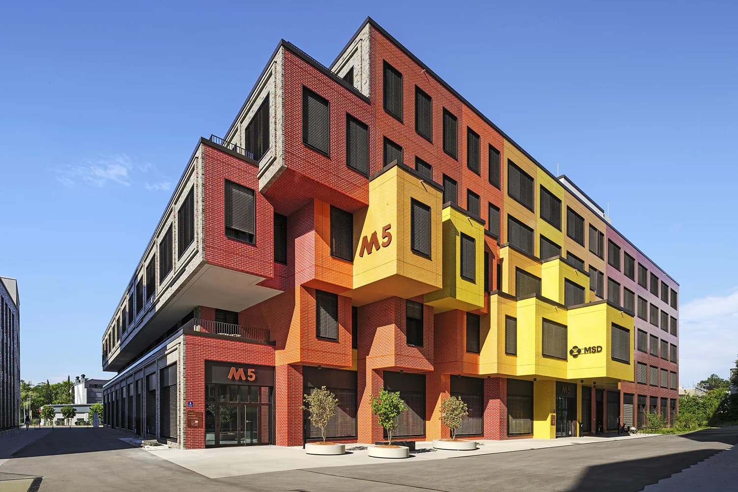 Vibrant mixed-use building with bold red, orange, and yellow façade featuring stacked geometric forms and large storefront windows, photographed under clear sky by Jessica Knobloch-Arscott.