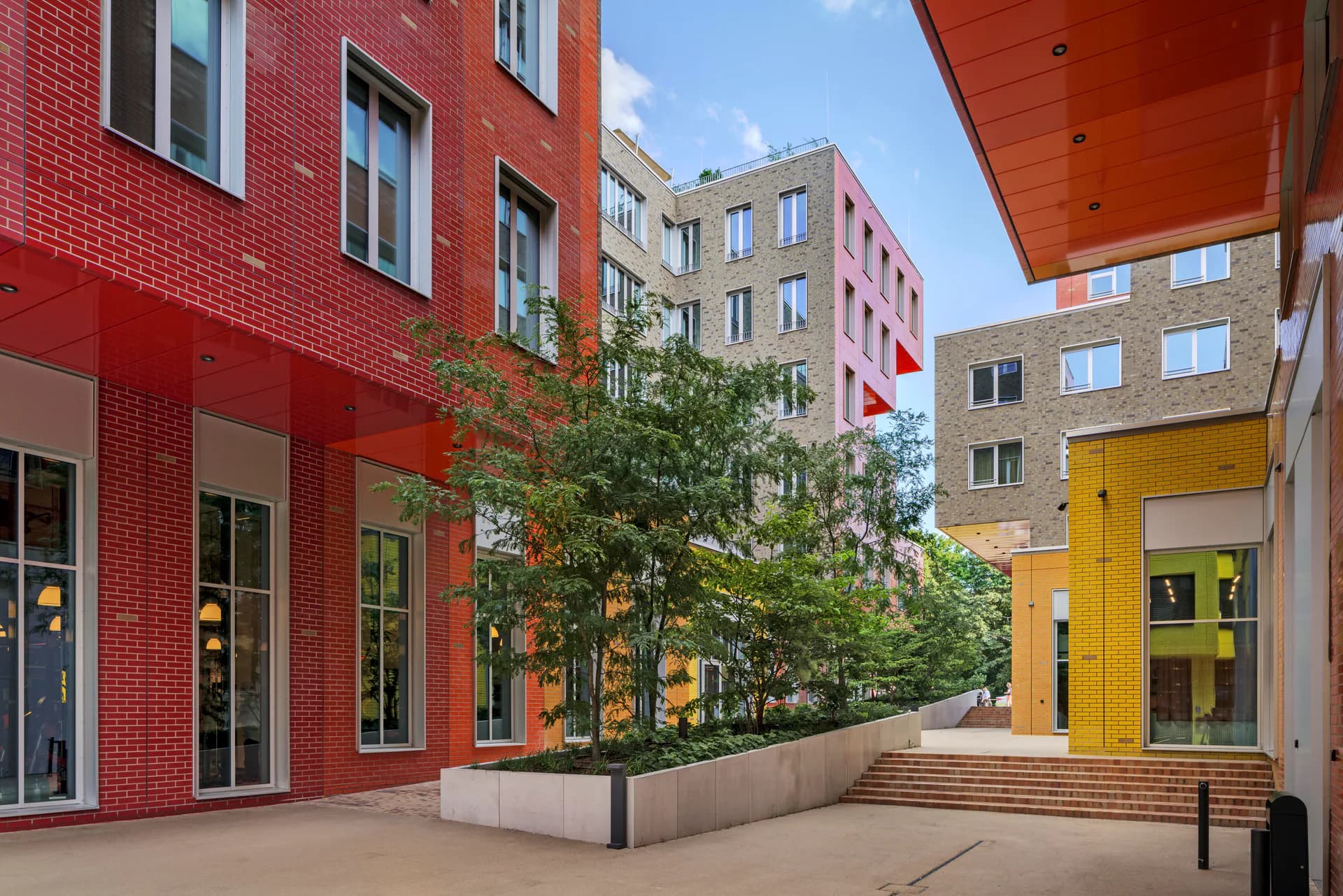 Vibrant courtyard with red brick and yellow buildings featuring modern architecture, green trees, and open plaza by Jessica Knobloch-Arscott.
