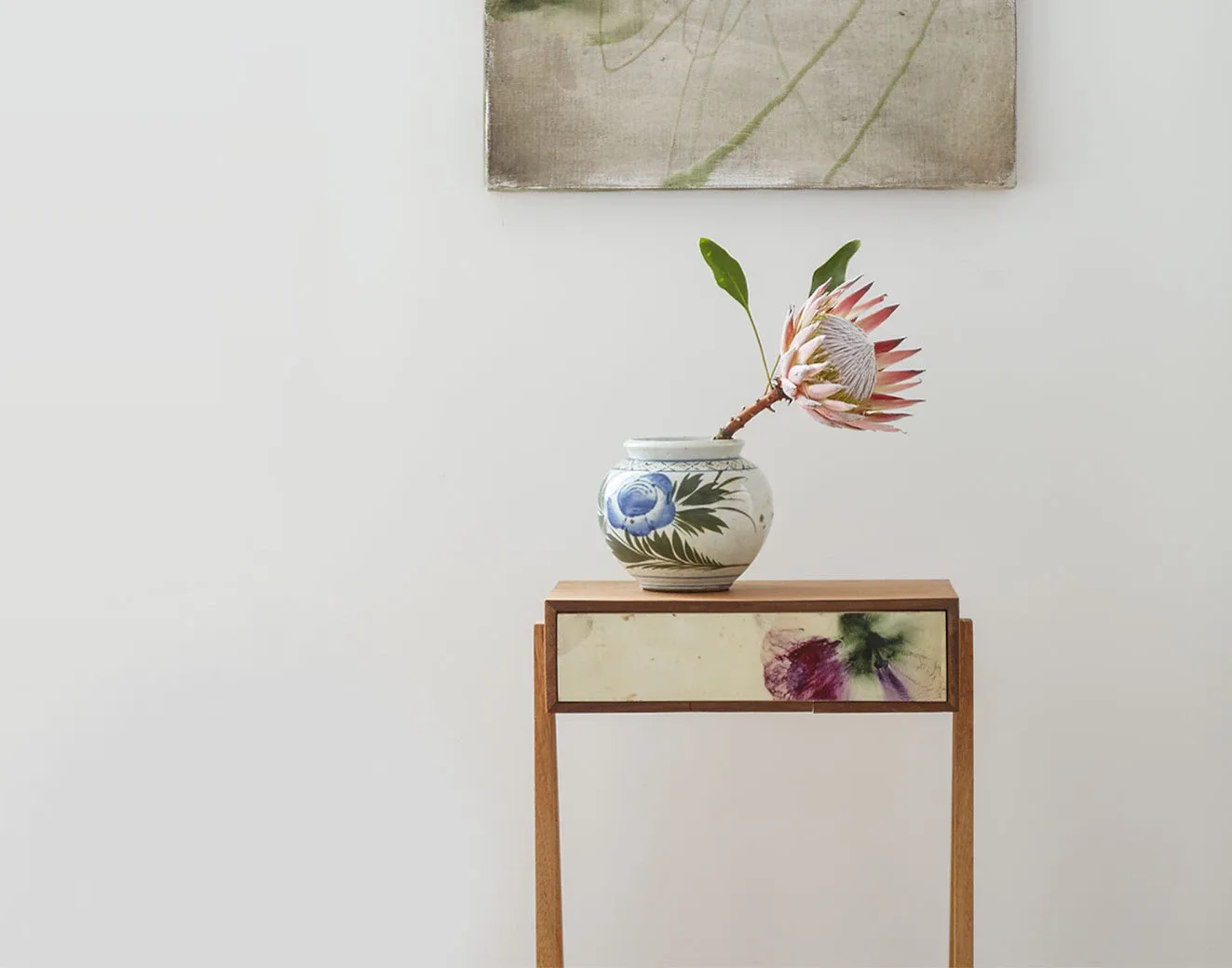 Minimalist interior styling featuring a wooden side table with decorative panel, ceramic vase with blue motif, and coral protea stem, against white wall with artwork above by Jessica Law.
