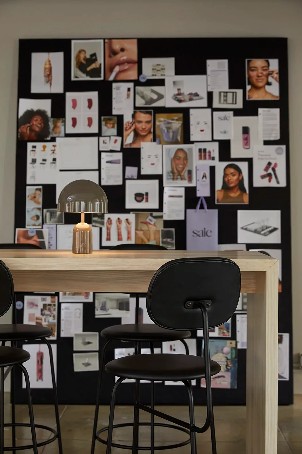 Black and white mood board wall installation above a natural wood desk with black chairs, featuring beauty product photography, makeup swatches, and brand materials by Jessica Werner.