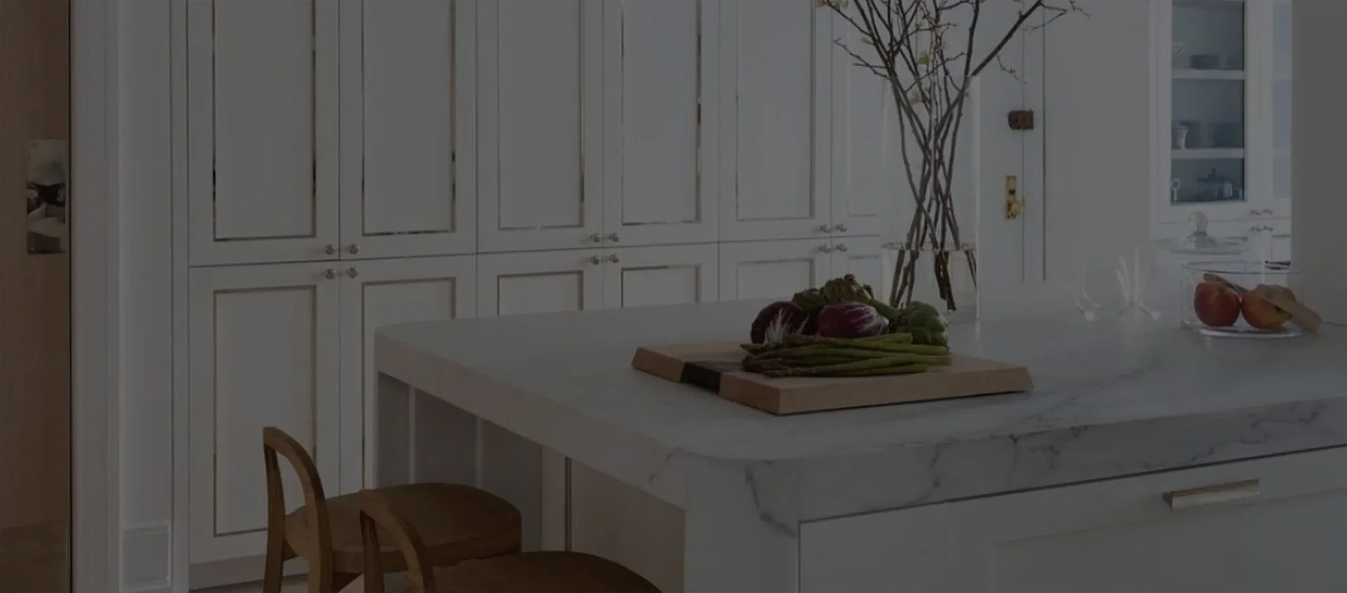 Modern kitchen with white cabinetry, gray countertop, and minimalist styling featuring a wooden chair and decorative vase by Jessie Childs.