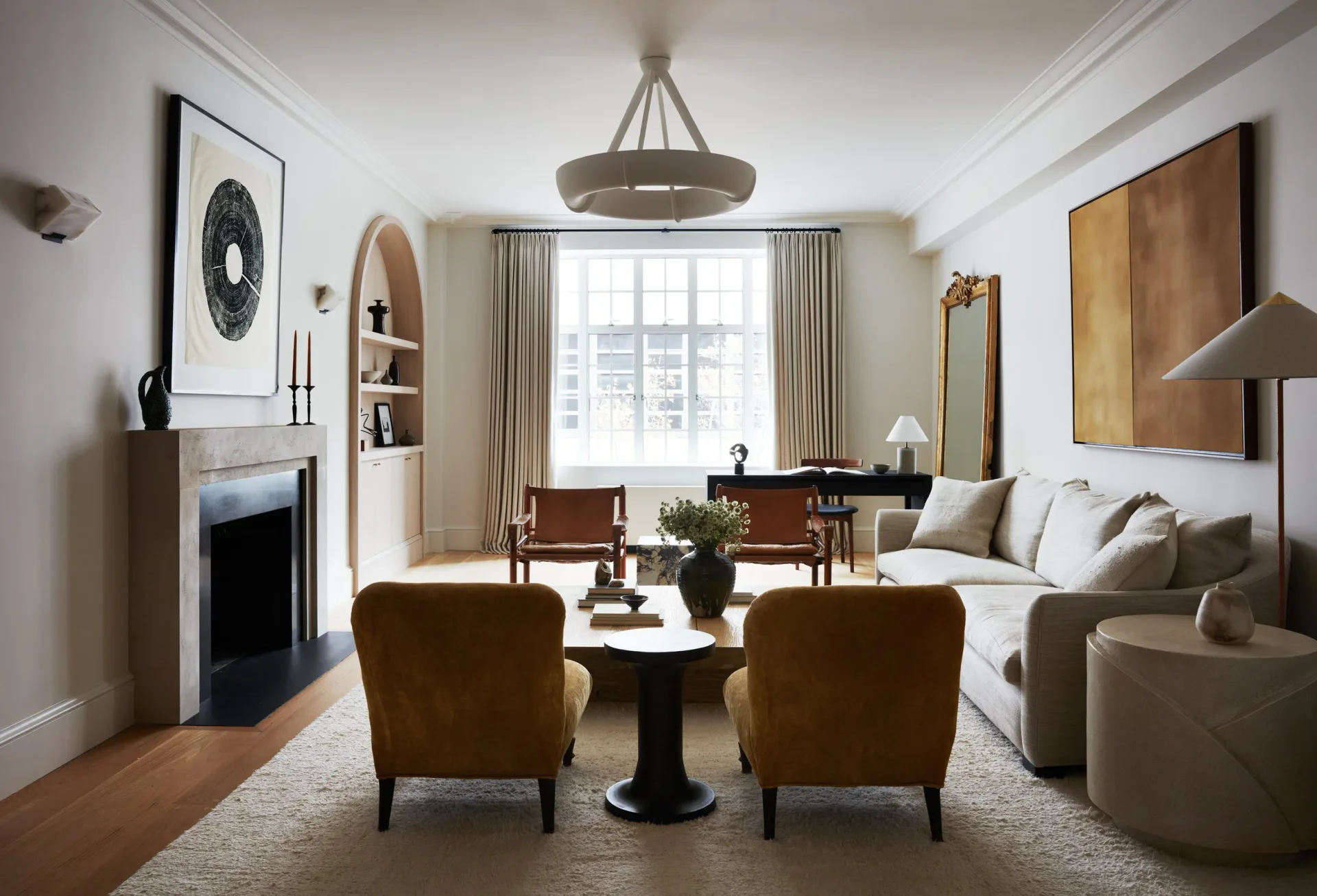 Elegant living room with cream sofa, cognac leather chairs, black pedestal table, fireplace, and large windows with natural light by Jessie Childs.
