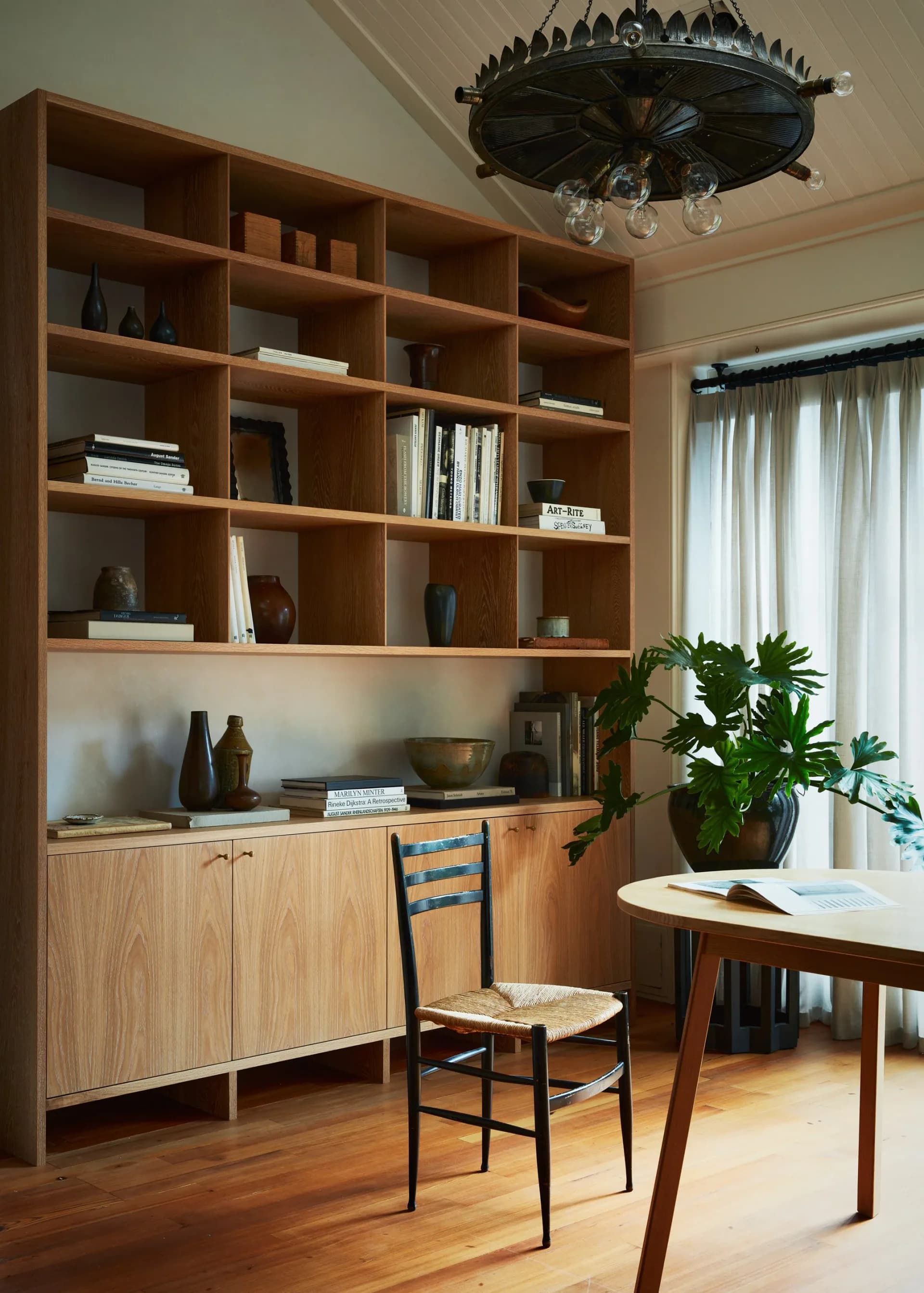 Modern home office with oak built-in shelving and cabinetry, round dining table, potted plant, and natural light from sheer-draped window by Jessie Childs.